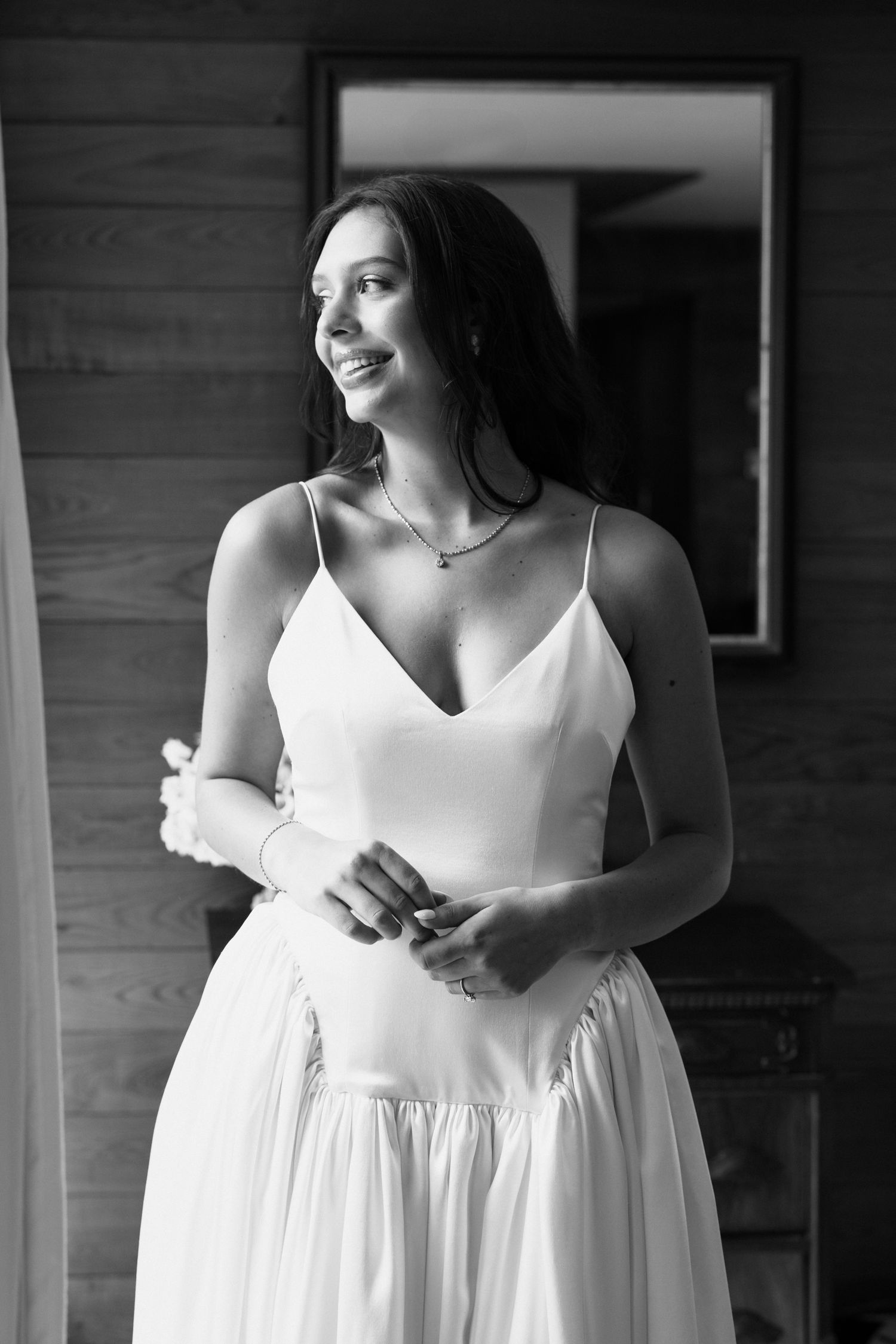 Black and white photo of a smiling bride in a white spaghetti strap wedding gown, standing indoors.