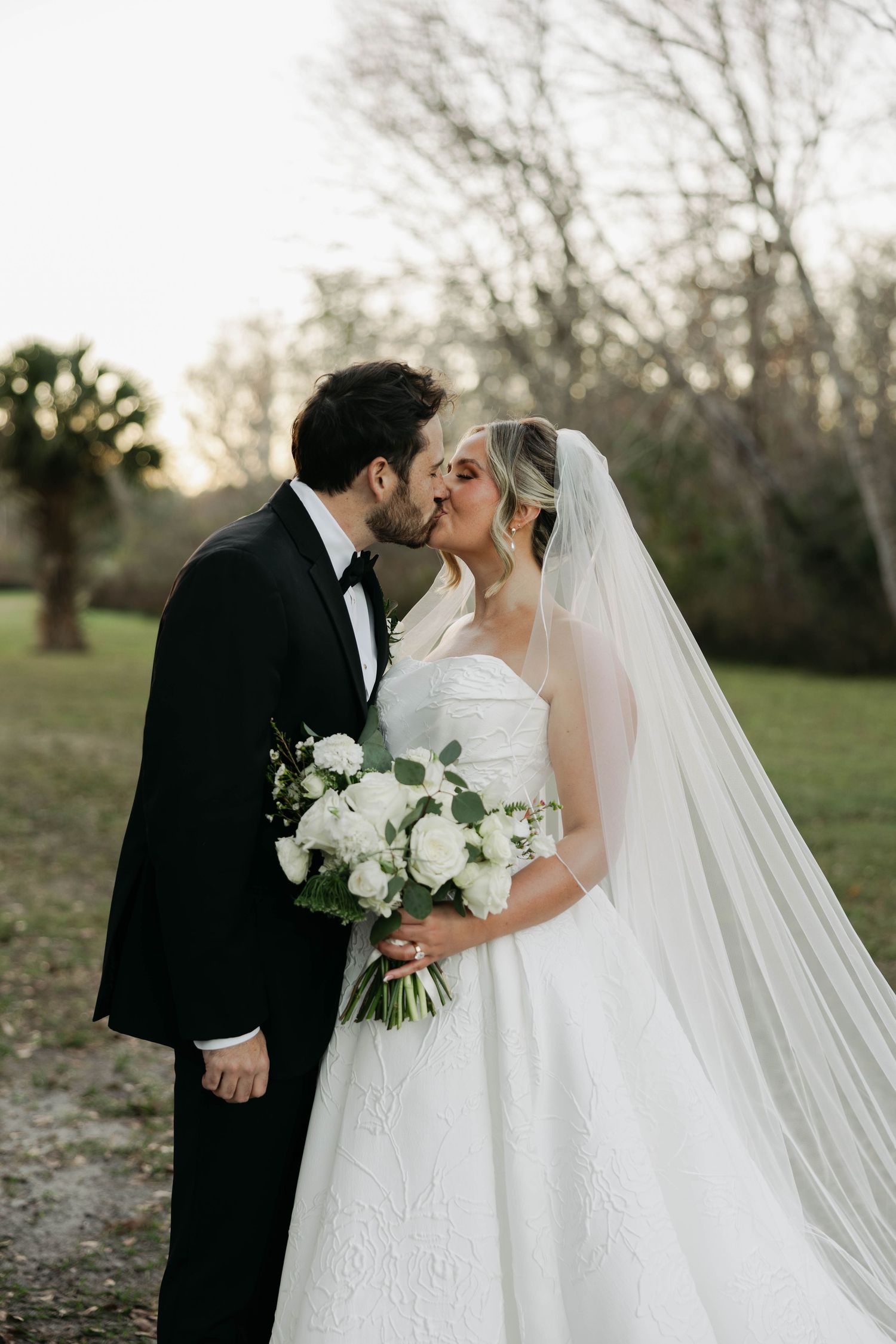Bride and groom sharing romantic kiss outdoors, bride holding white rose bouquet with long cathedral veil.