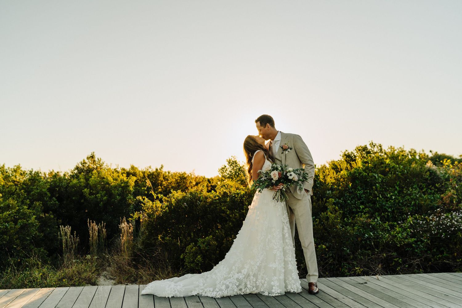 Bride and groom share a romantic sunset kiss on a wooden deck surrounded by lush greenery in wedding attire.