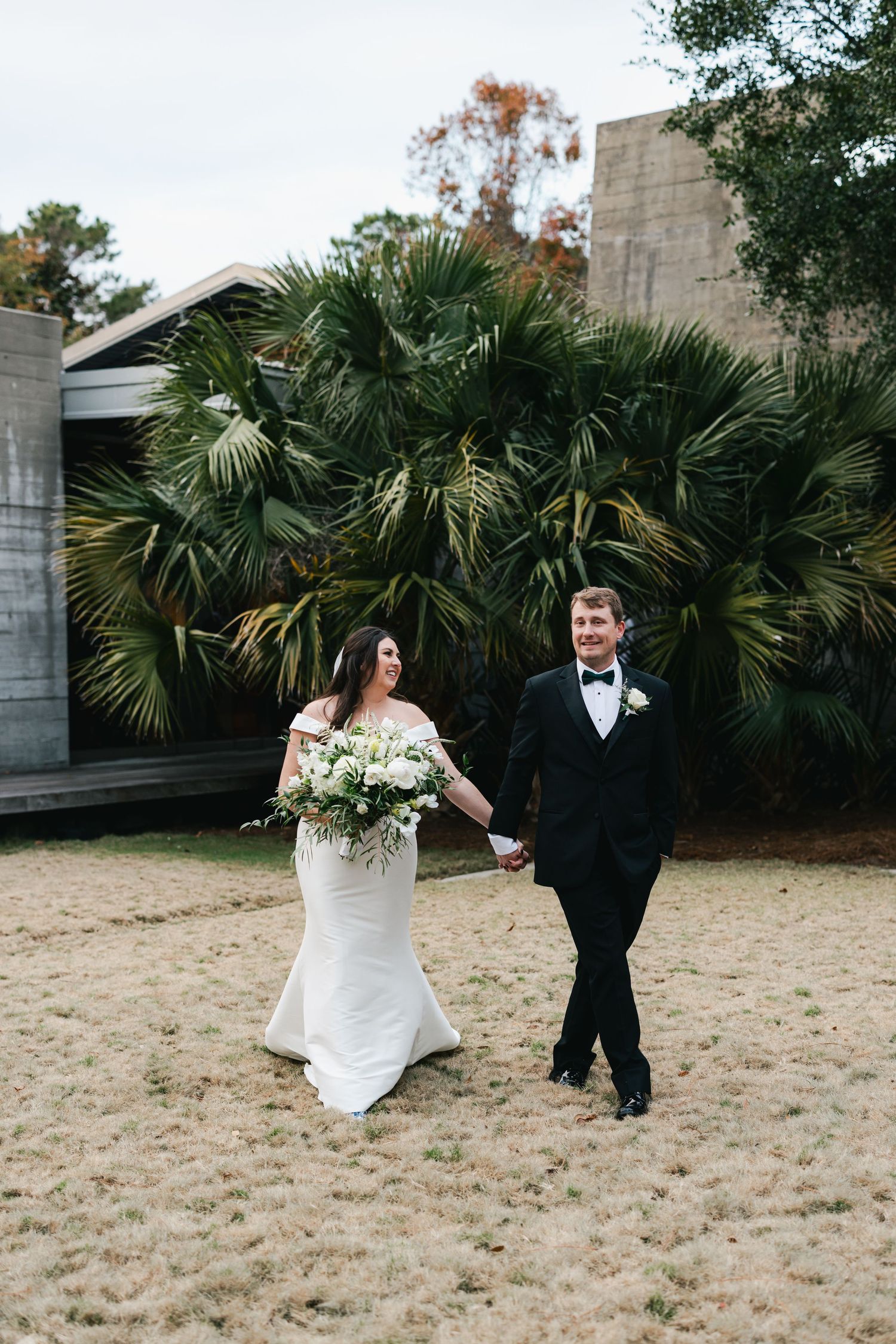 Bride and groom walk hand-in-hand outdoors, she in a white gown with bouquet, he in a black tuxedo, palm trees behind.