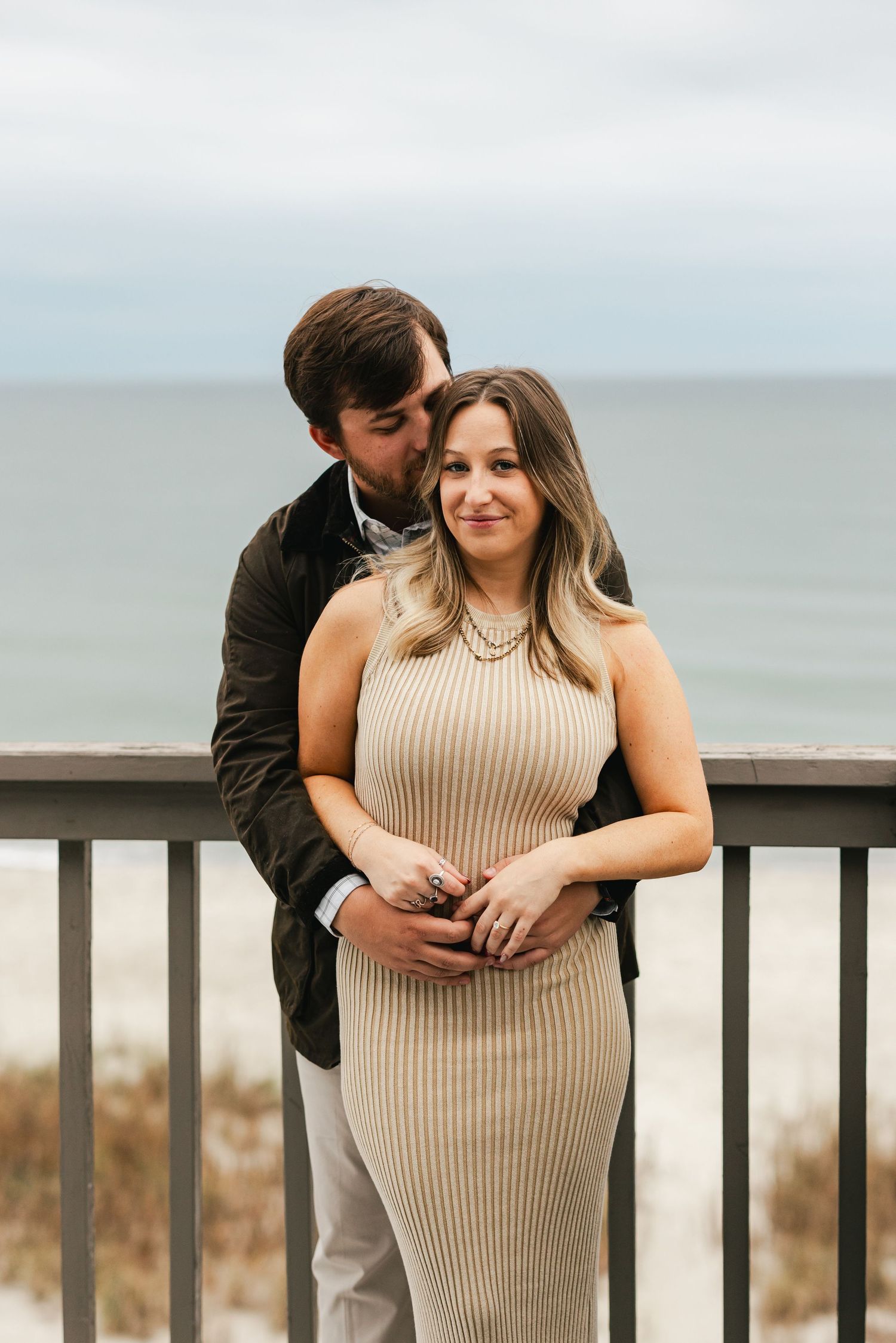 Couple embracing on a beach balcony, man kissing woman's head, ocean backdrop, she wears a ribbed beige dress.