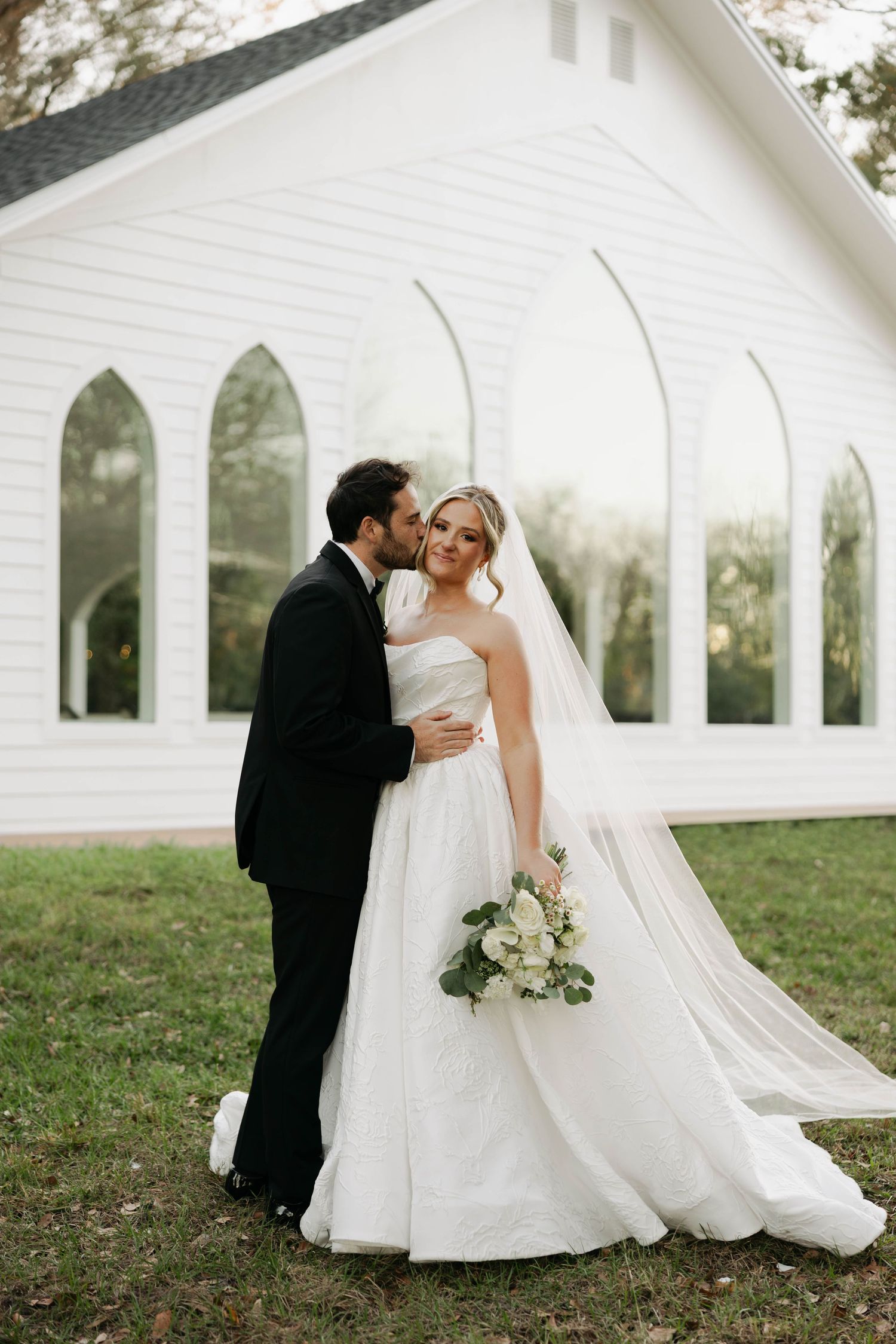 Bride and groom embrace outside a white chapel, bride in a flowing gown with veil and bouquet of white roses.