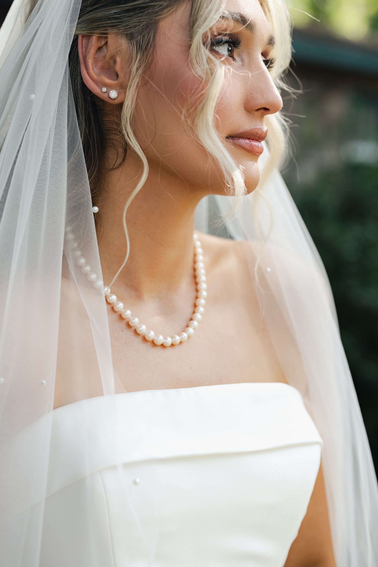 Close-up of bride wearing pearl necklace and earrings with white veil and strapless wedding gown outdoors.