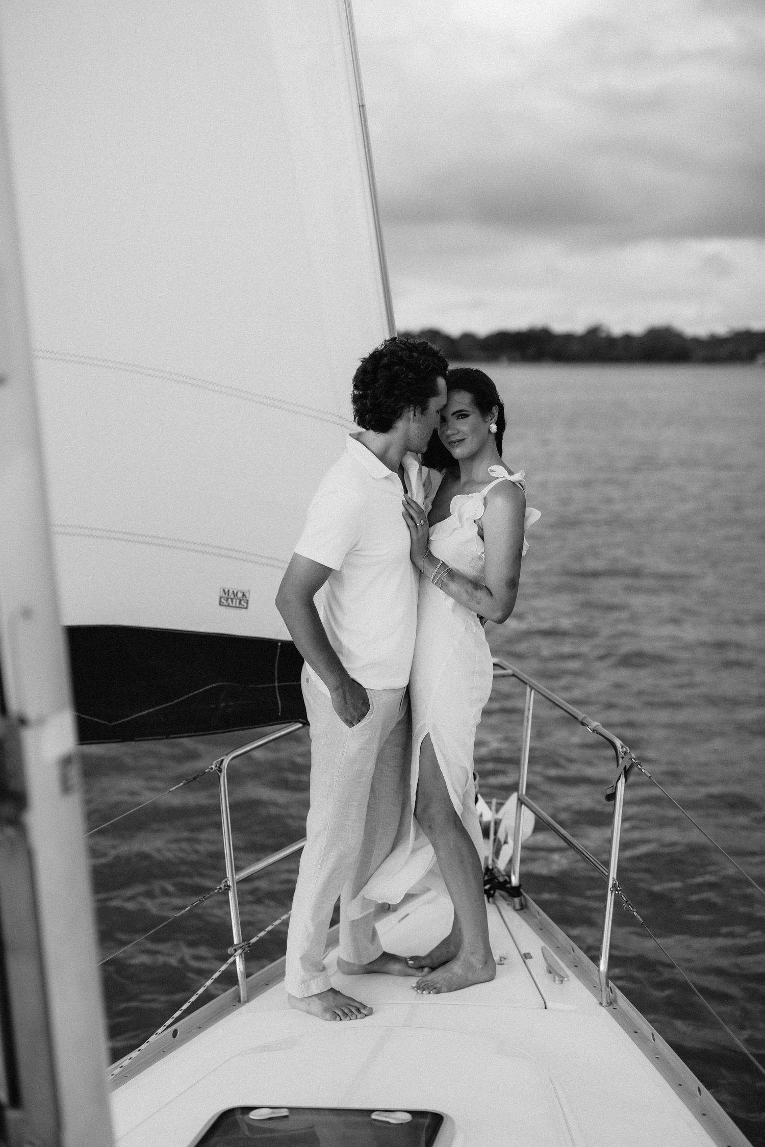 Black and white photo of a couple embracing on the bow of a sailboat on open water.