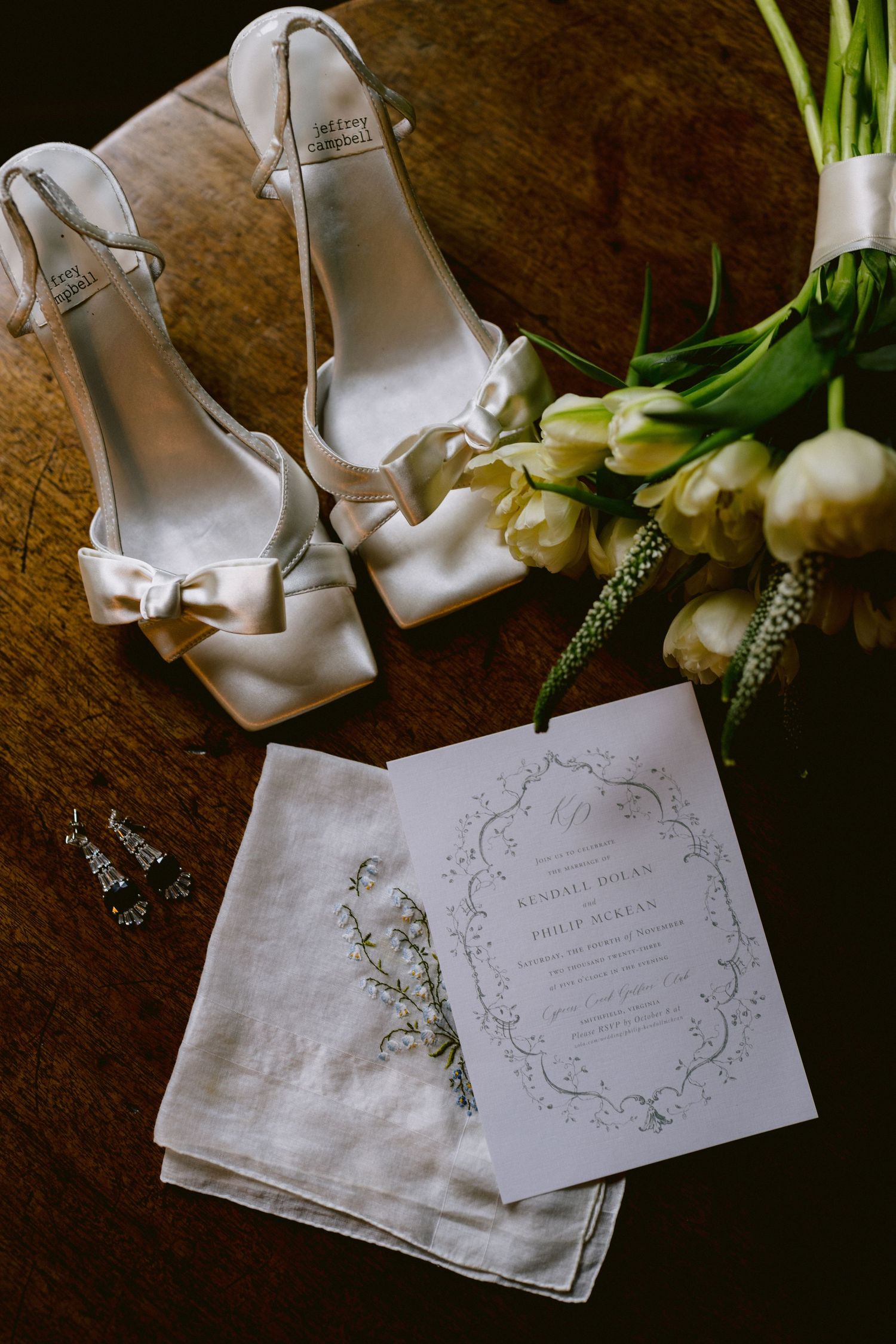 Flat lay of white satin bow heels, wedding invitation, tulip bouquet, and drop earrings on wooden surface.