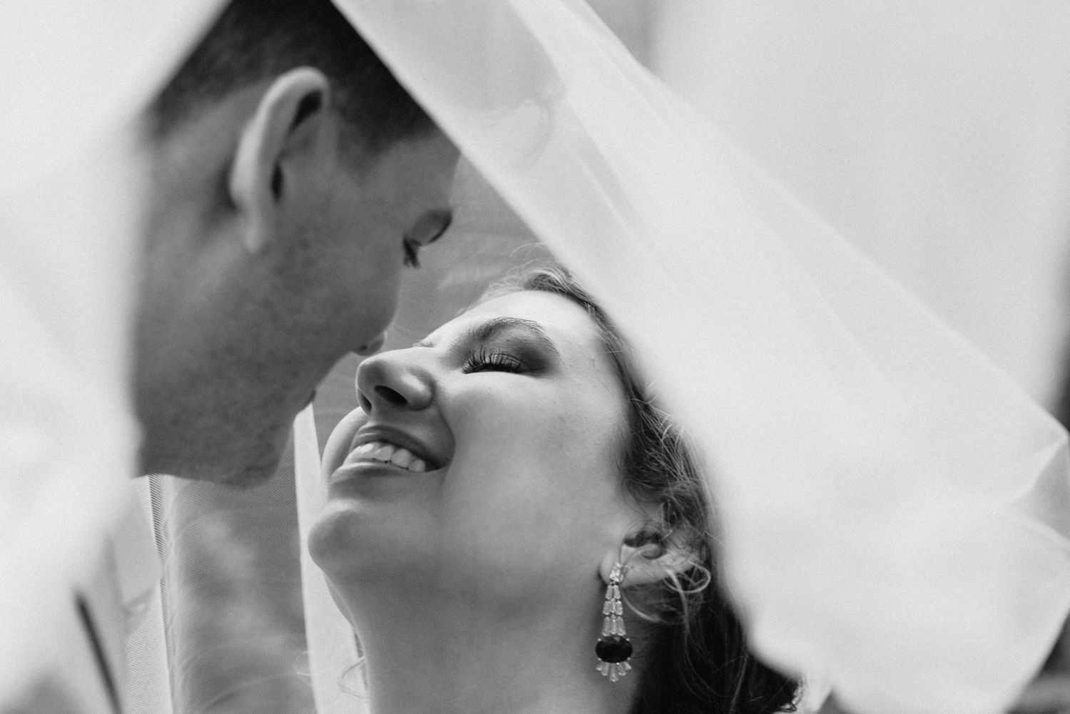 Black and white romantic close-up of smiling bride and groom nearly kissing under flowing veil.