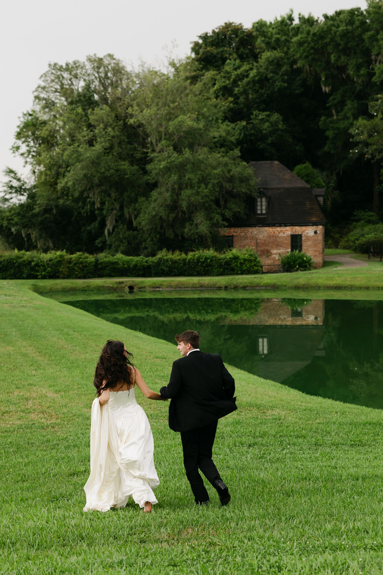 Bride and groom walking hand in hand across lush green lawn beside a reflective pond with historic brick building backdrop.