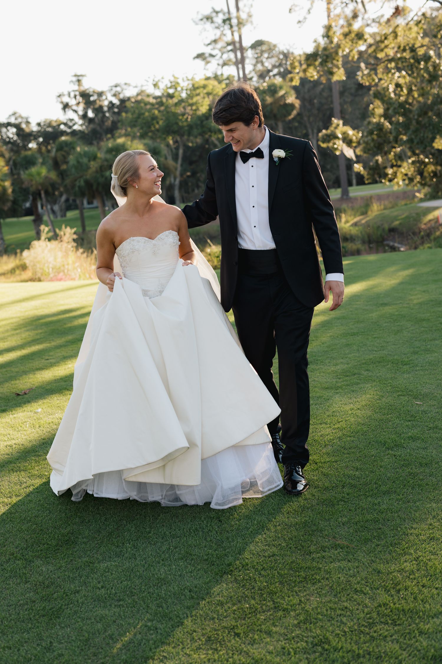 Bride and groom walking on green grass, woman in white strapless gown, man in black tuxedo with boutonniere.