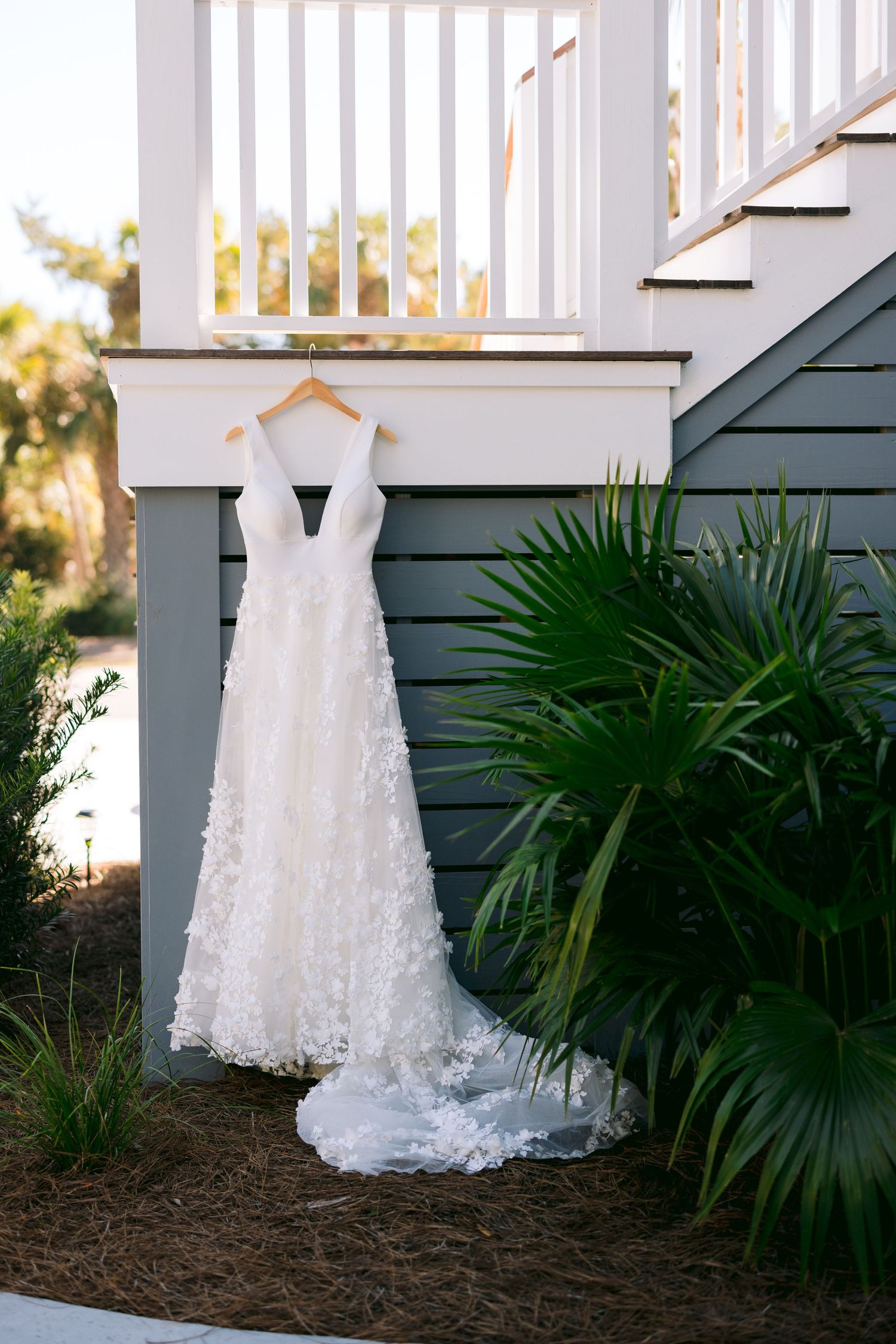 White floral A-line wedding dress hanging on wooden hanger at base of outdoor staircase with tropical greenery.