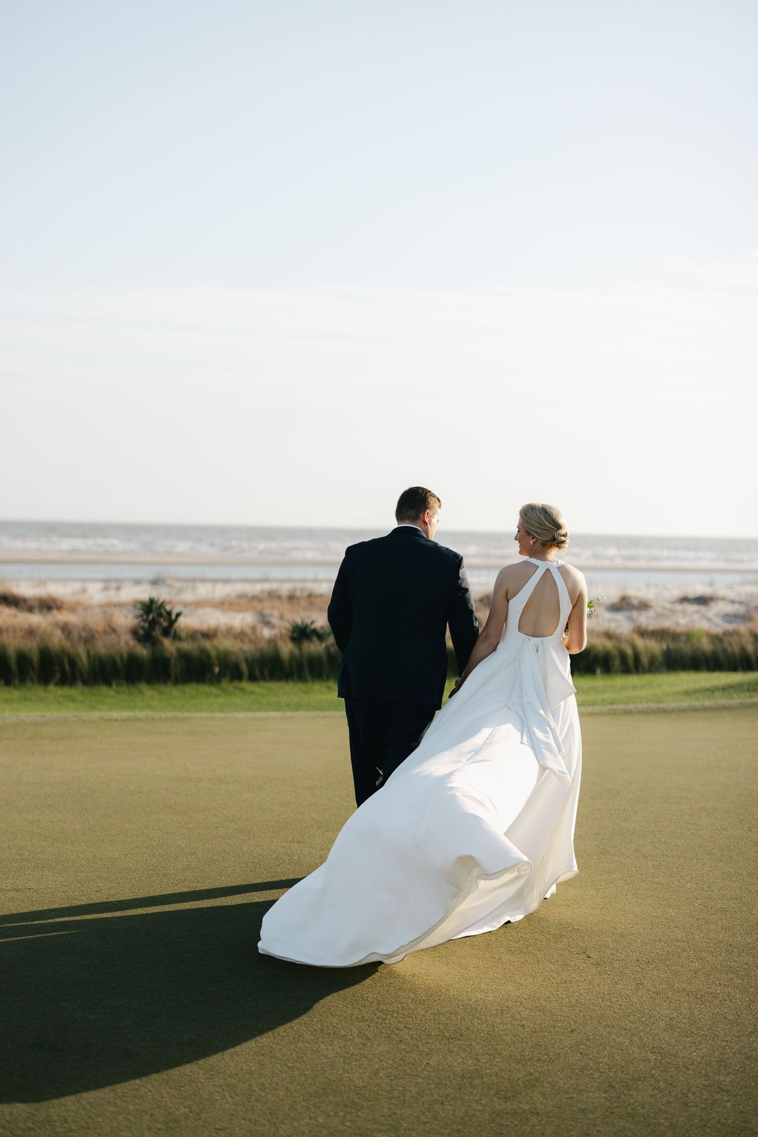 Bride and groom walking hand in hand toward the ocean on a golf course, wedding dress trailing in the breeze.