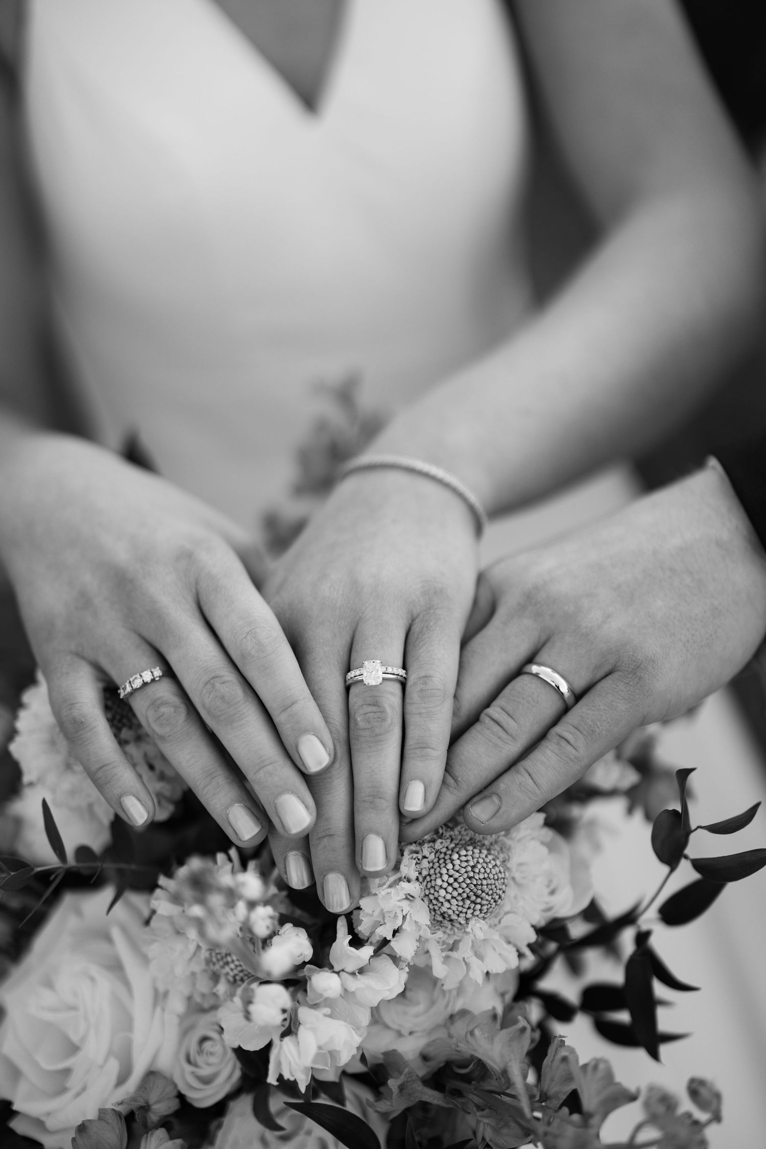 Black and white photo of three hands displaying wedding and engagement rings resting on a bridal bouquet.