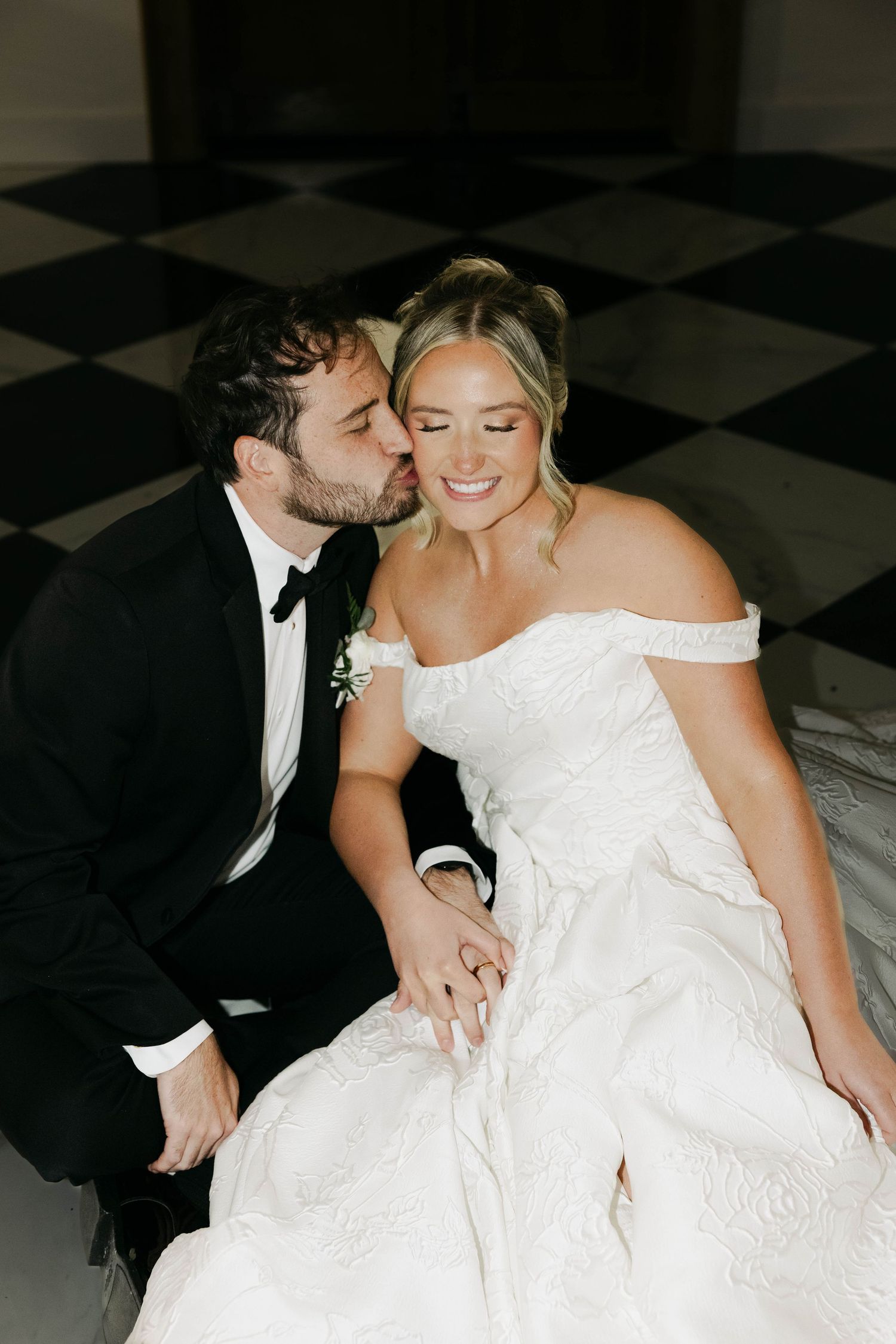 Groom in tuxedo kissing bride in off-shoulder gown on black and white checkered floor.