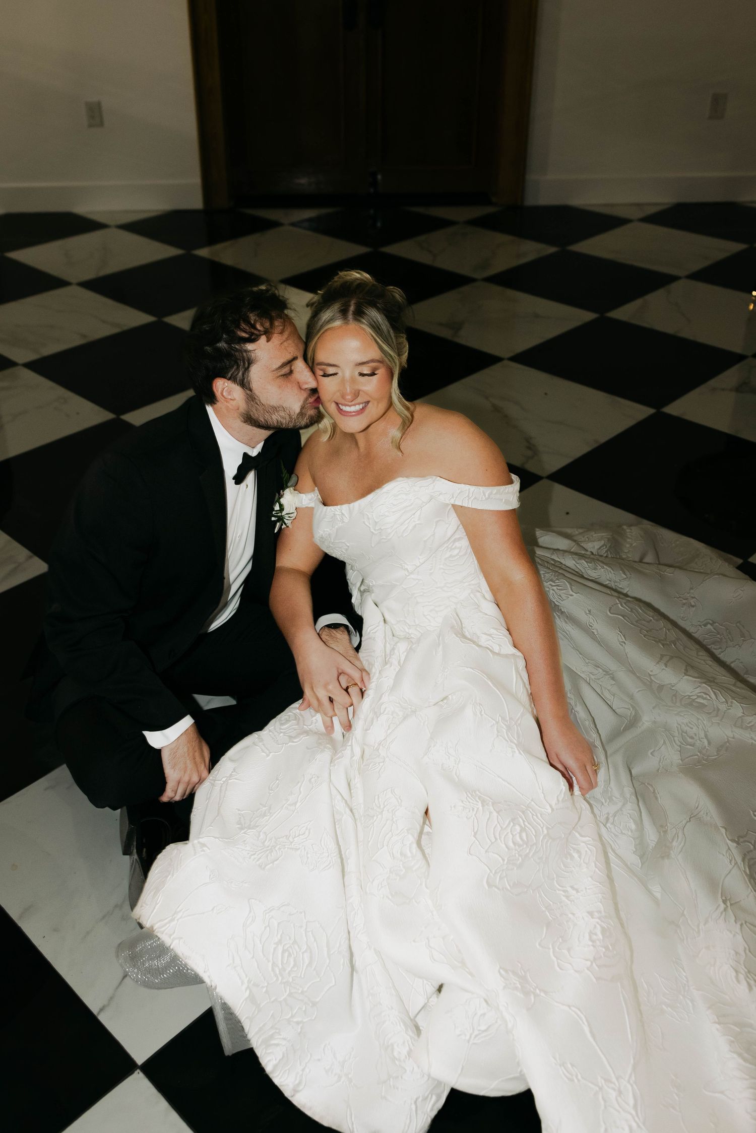 Bride and groom sitting on black and white checkered floor, groom kissing bride's cheek in elegant ballroom.