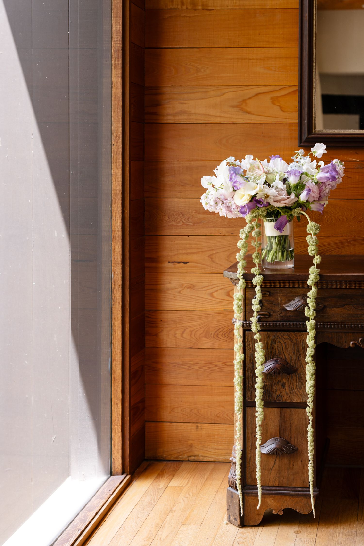 Elegant wedding bouquet with purple and white flowers in glass vase on antique wooden stand with trailing greenery.