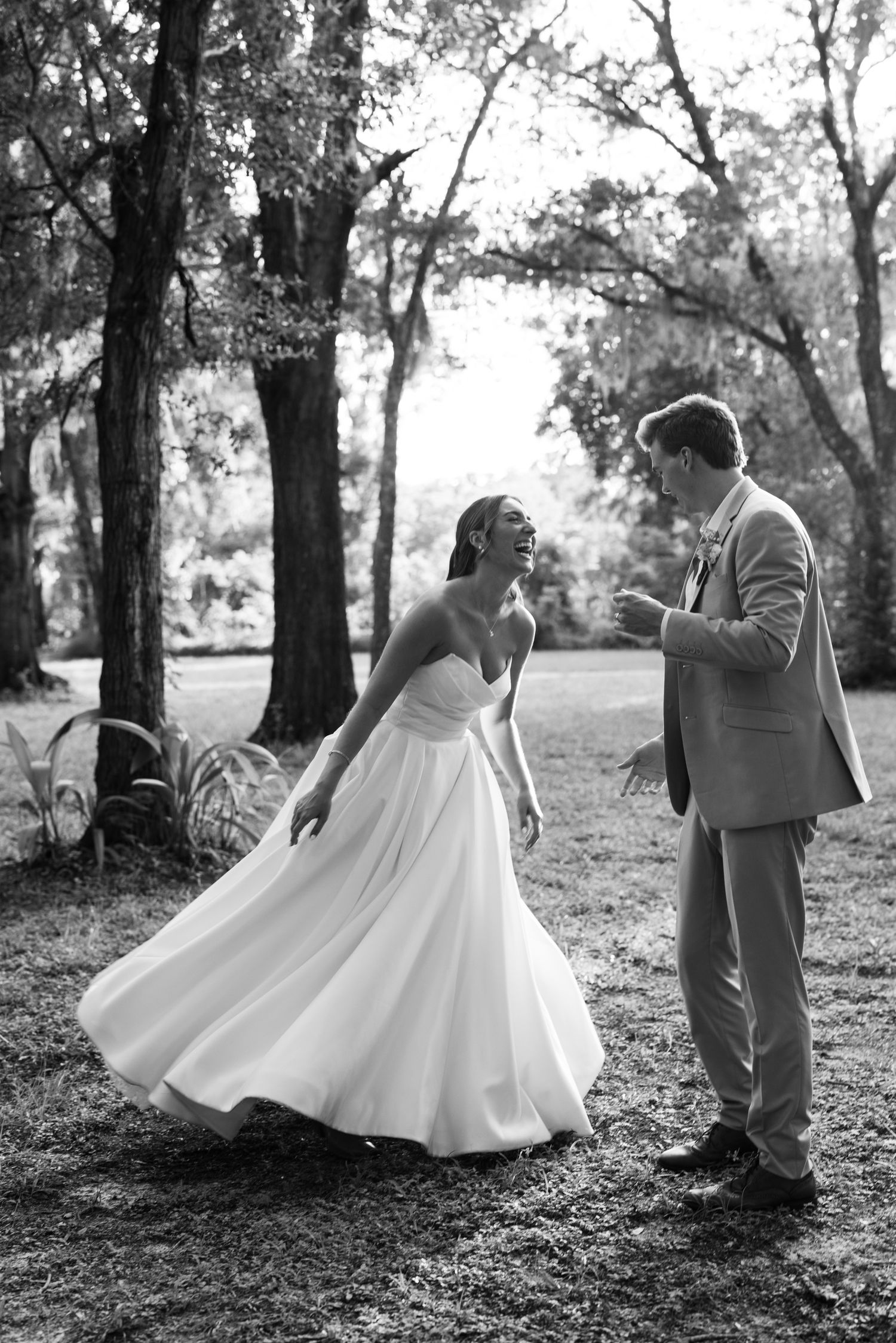 Black and white photo of a bride in a flowing gown twirling for her groom in a suit outdoors among trees.
