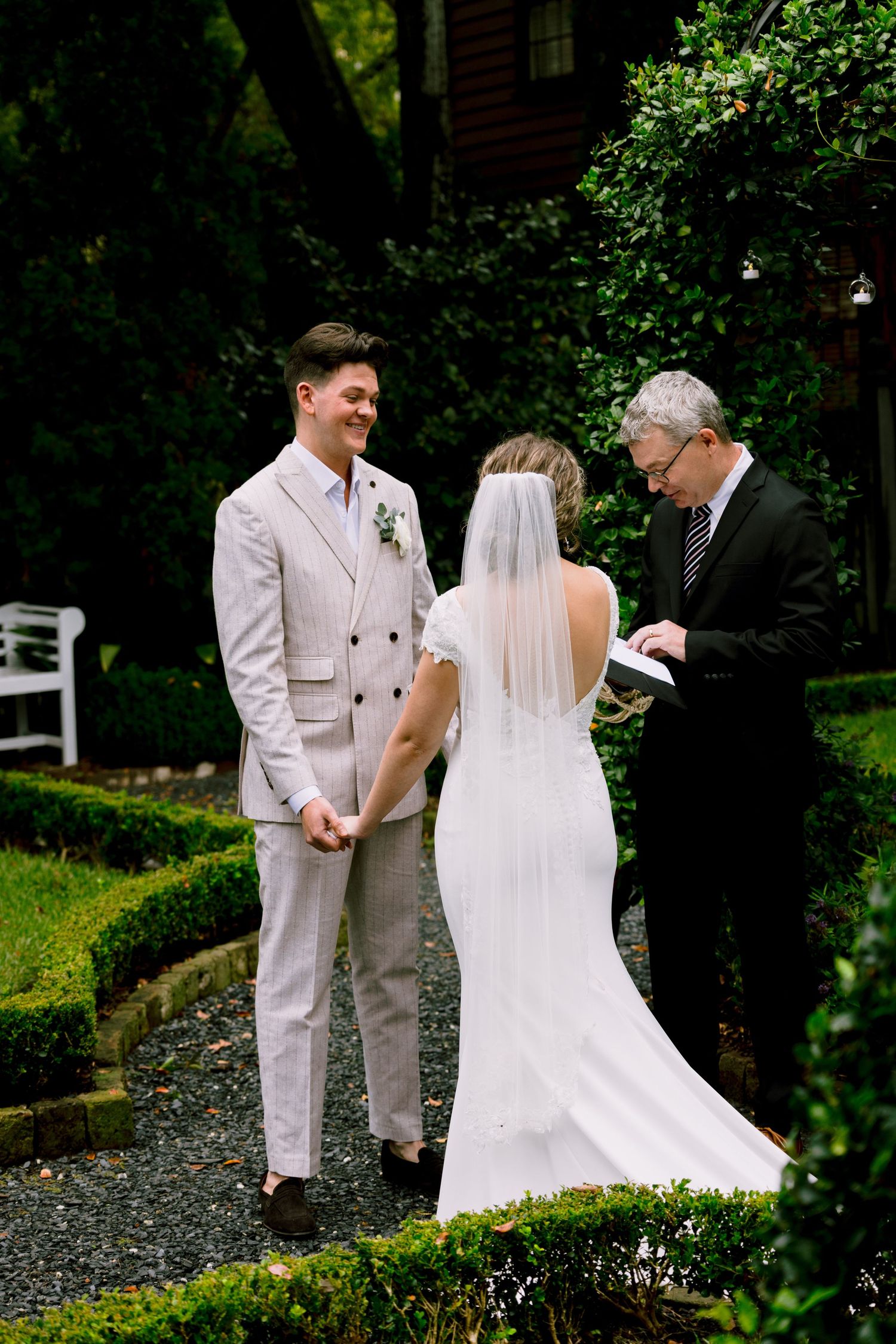 Outdoor garden wedding ceremony with smiling groom in beige suit and bride in white gown with veil.
