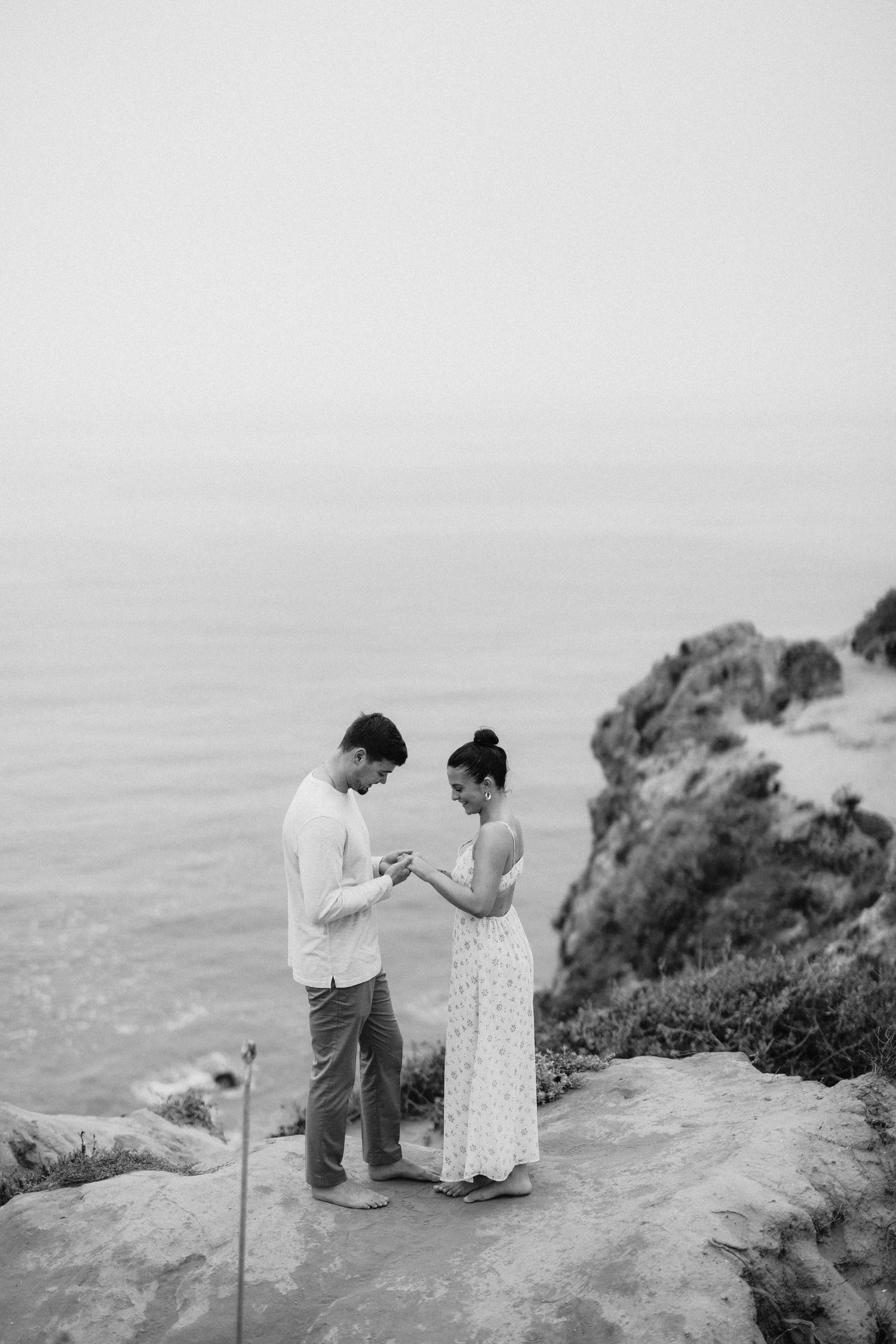 Black and white photo of couple sharing engagement ring moment on coastal cliff overlooking ocean.
