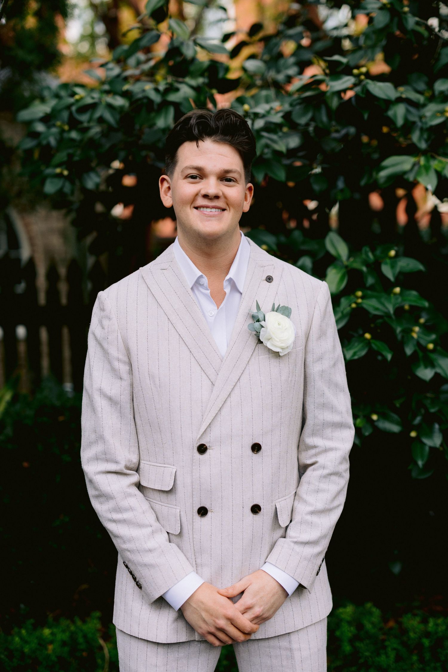 Smiling groom in pinstripe double-breasted suit with white rose boutonniere standing before green foliage.