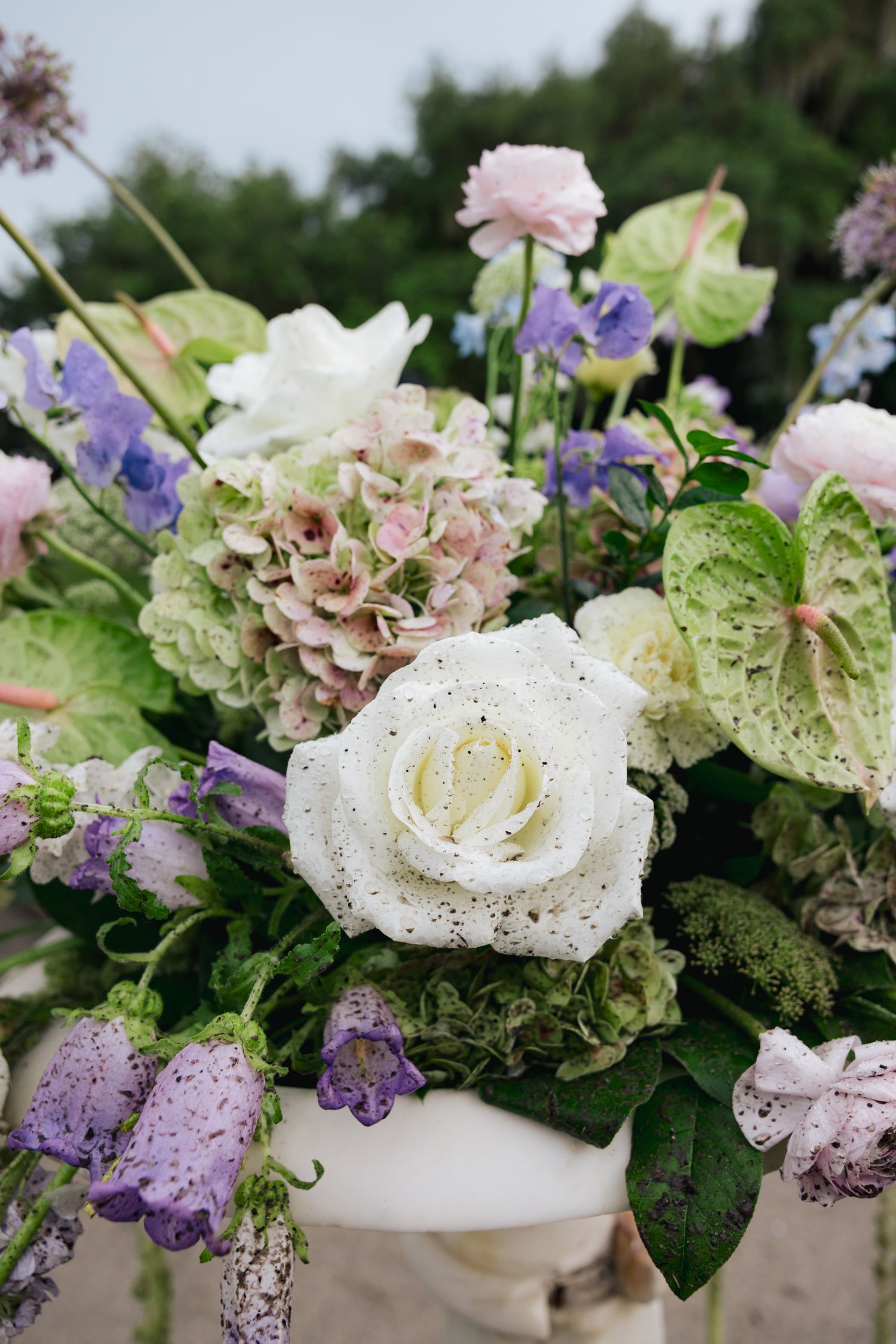 Elegant floral arrangement featuring a white rose, purple hydrangeas, lavender bells, and green anthurium leaves.