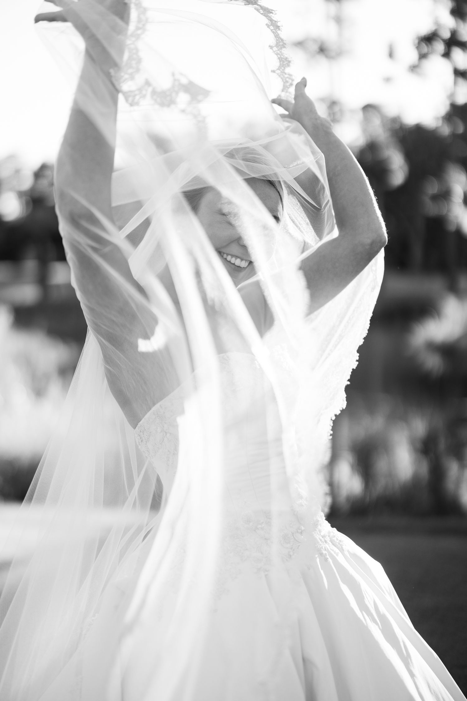 Black and white photo of a smiling bride tossing her flowing veil in the air outdoors.