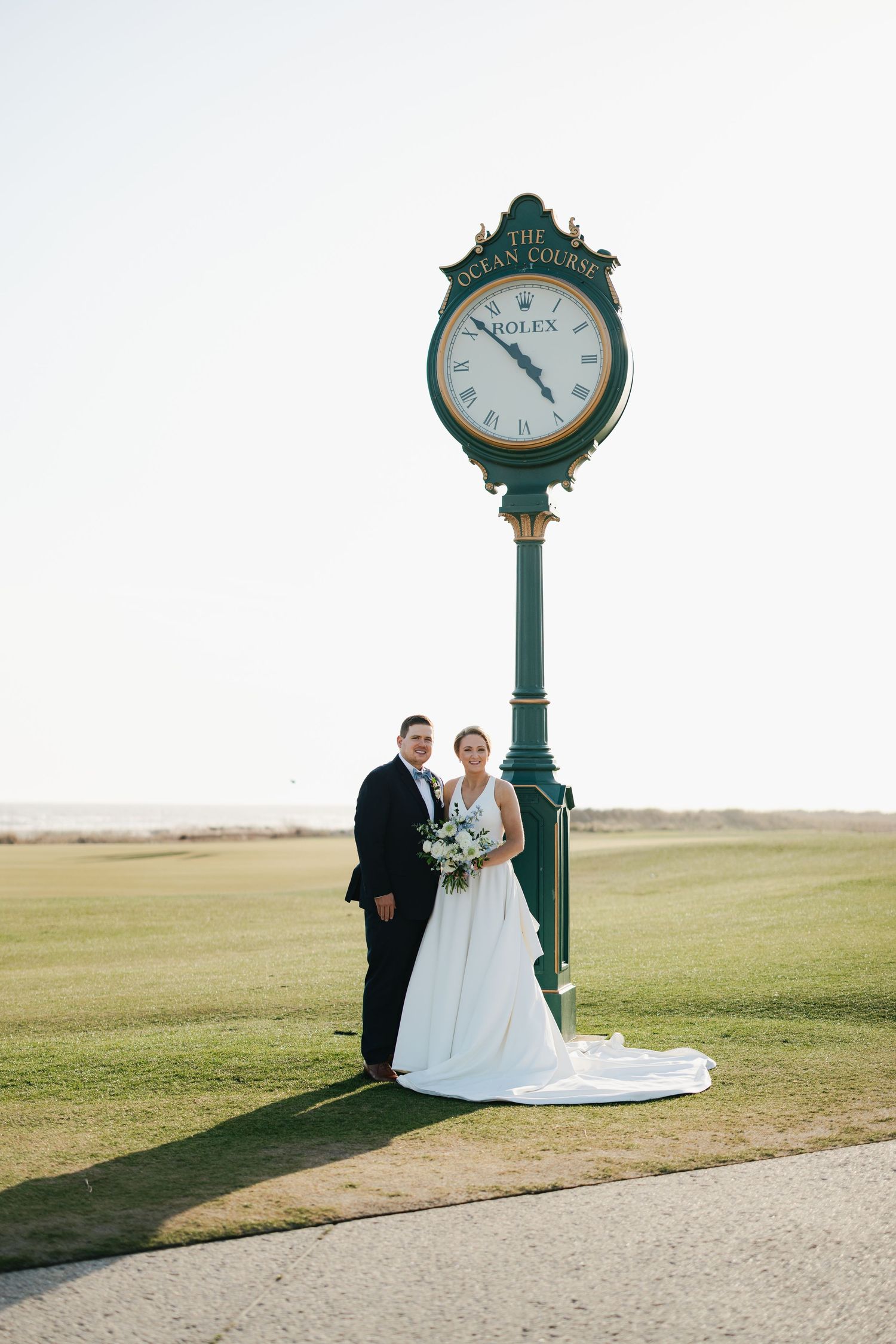 Bride and groom posing beside a Rolex clock at The Ocean Course golf club on their wedding day.