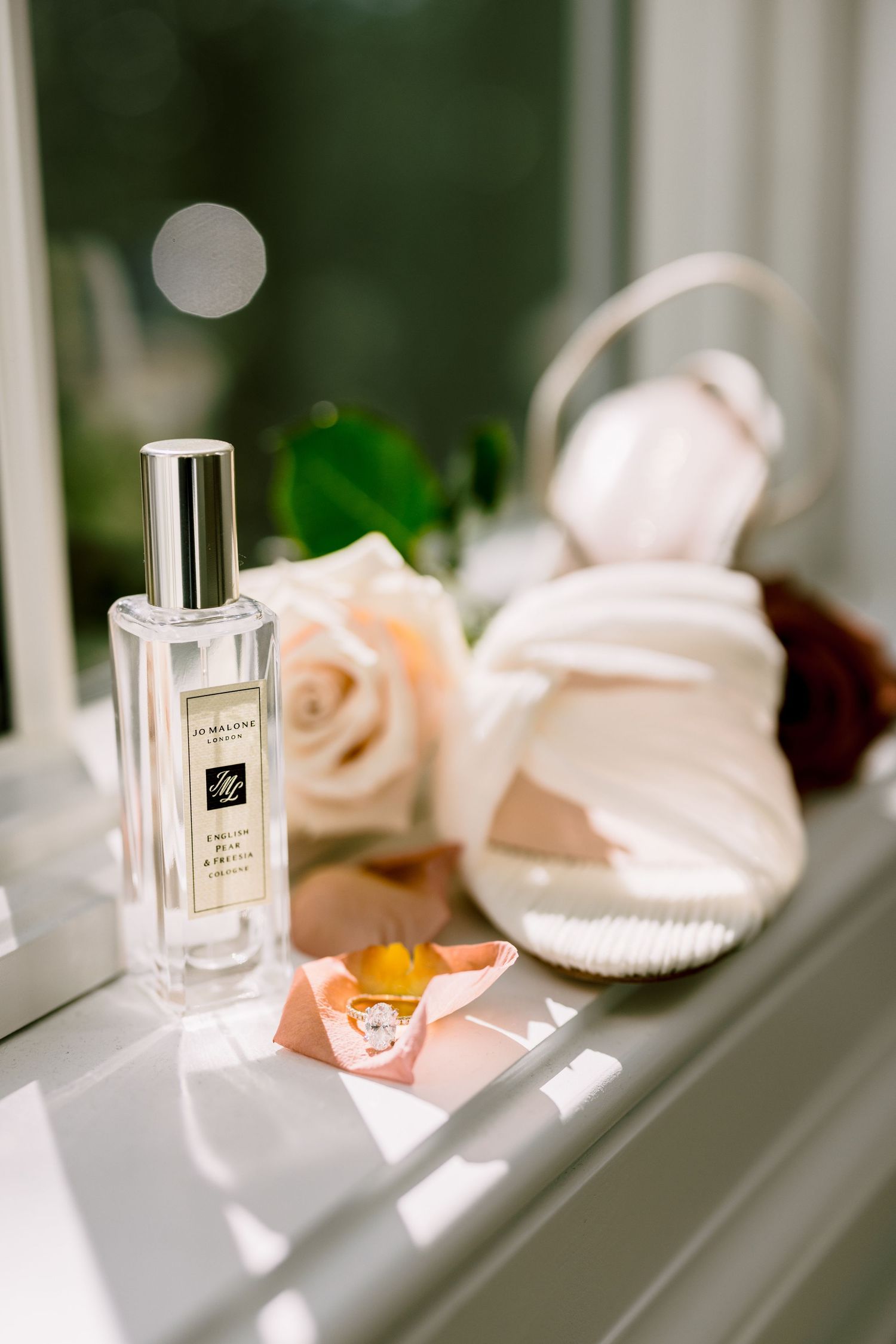 Jo Malone perfume bottle beside a white strappy heel, blush roses, and a diamond ring on a sunlit windowsill.