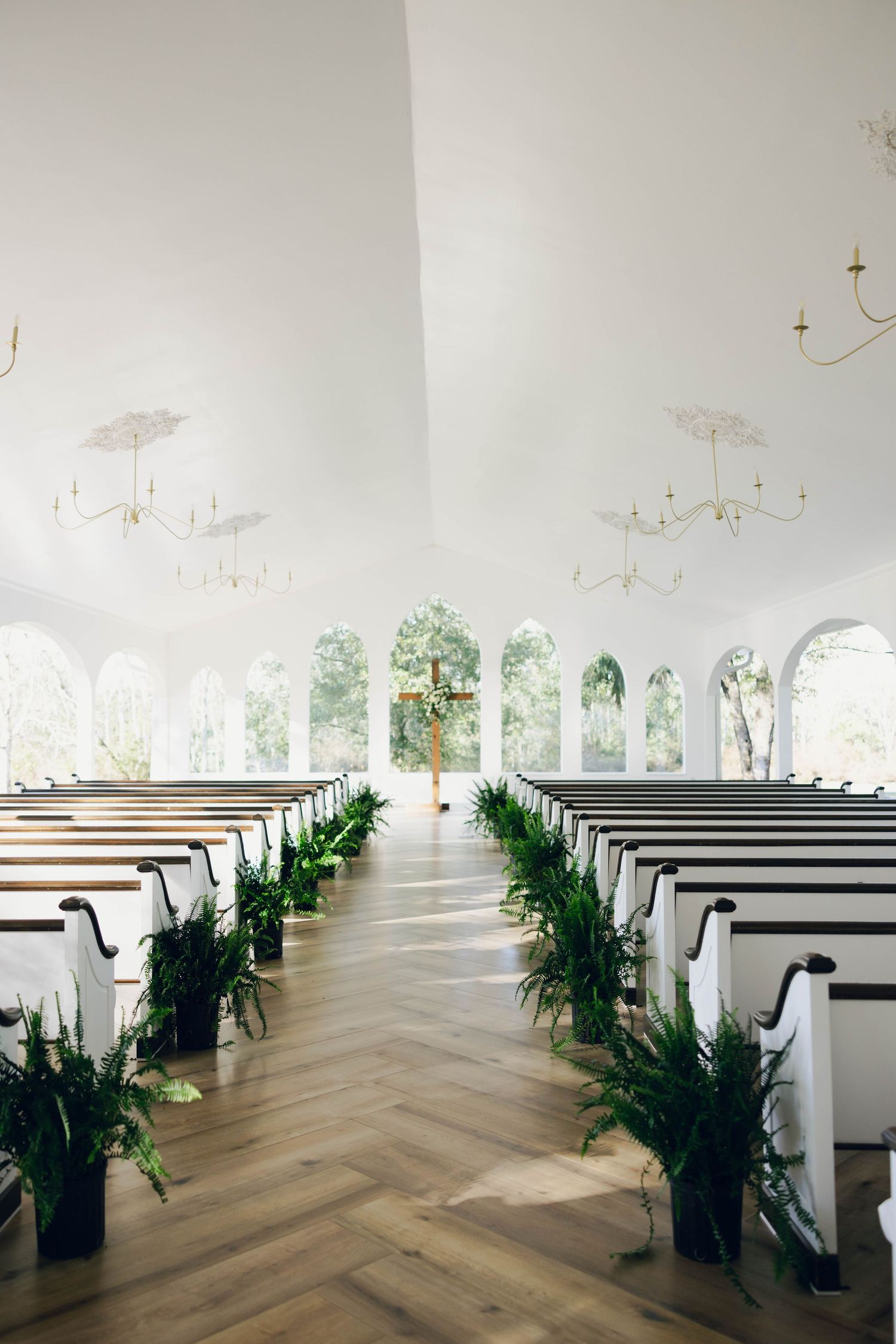 Elegant white wedding chapel interior with herringbone floors, white pews, greenery aisle decor, and gold chandeliers.
