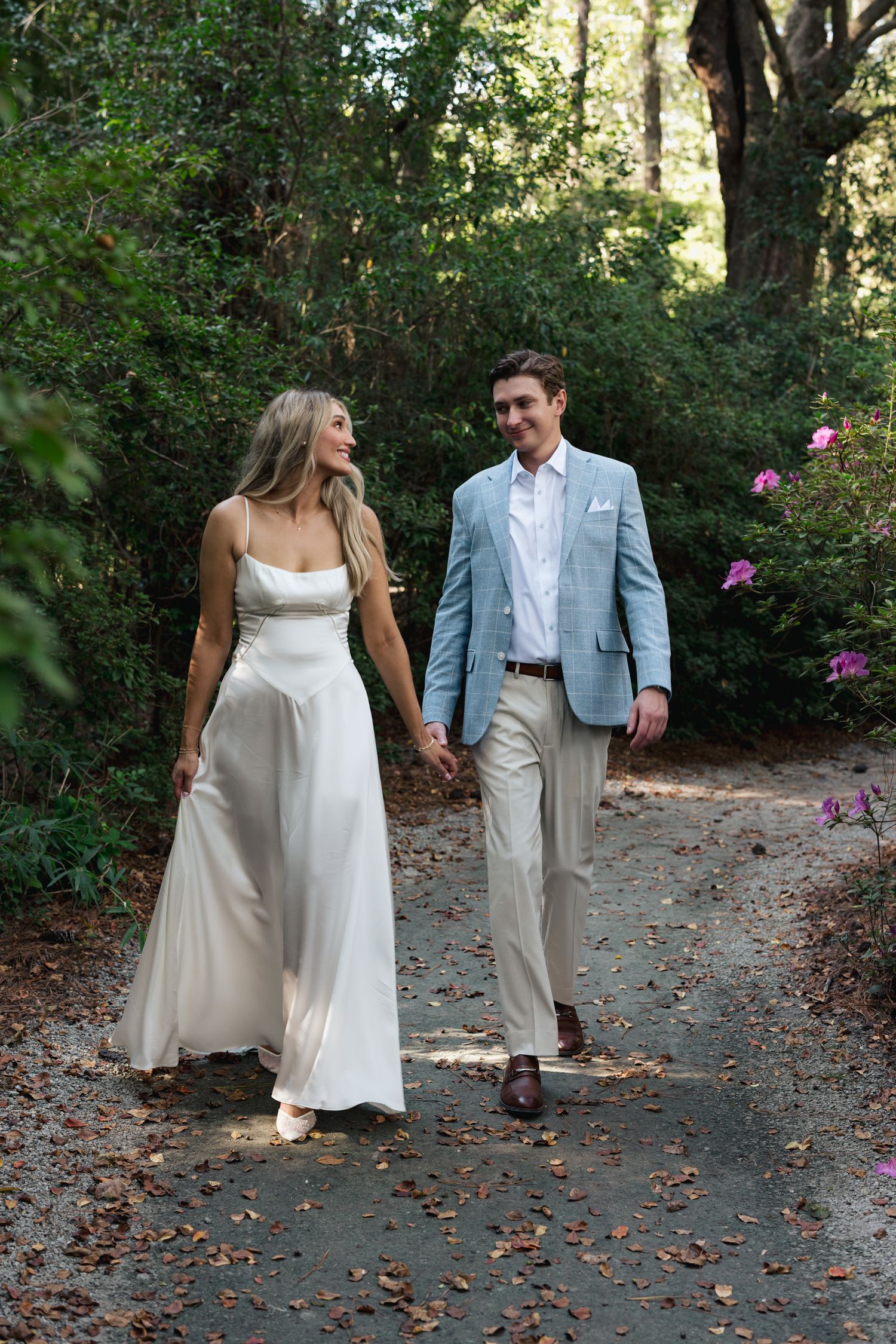 A bride in a white gown and groom in a light blue blazer walk hand in hand along a garden path.