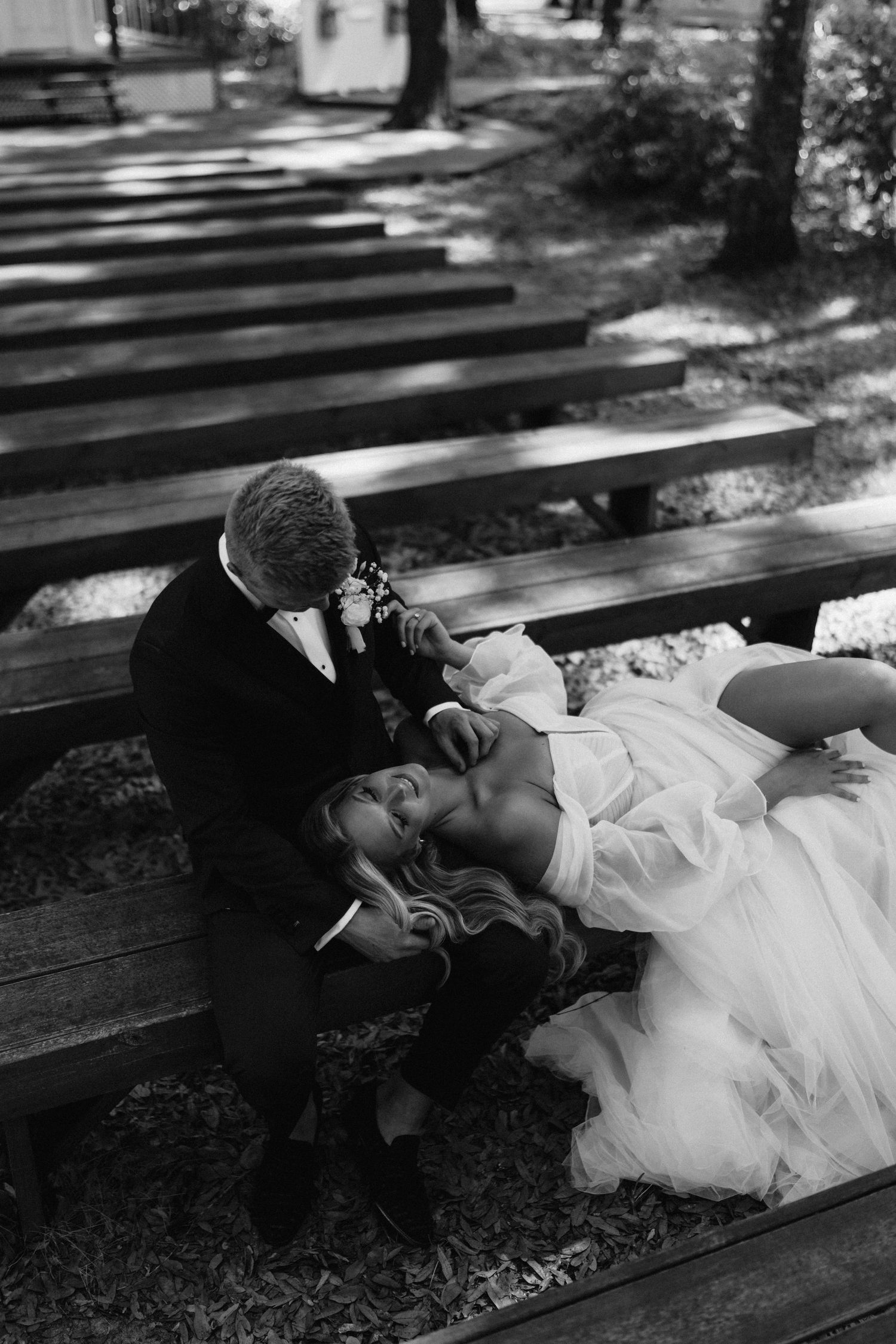 Black and white photo of bride lying on groom's lap on outdoor wooden bench at wedding ceremony.