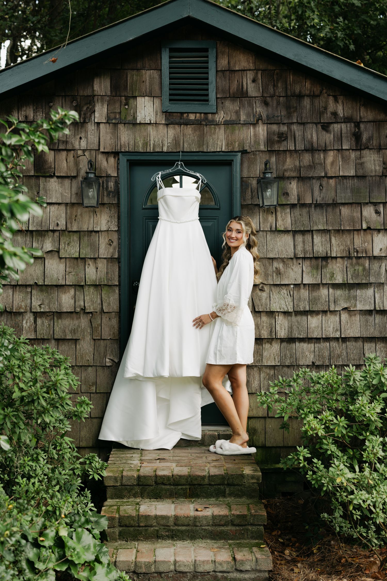 Smiling bride in a white robe poses beside her strapless wedding gown hanging on a rustic wooden chapel door.