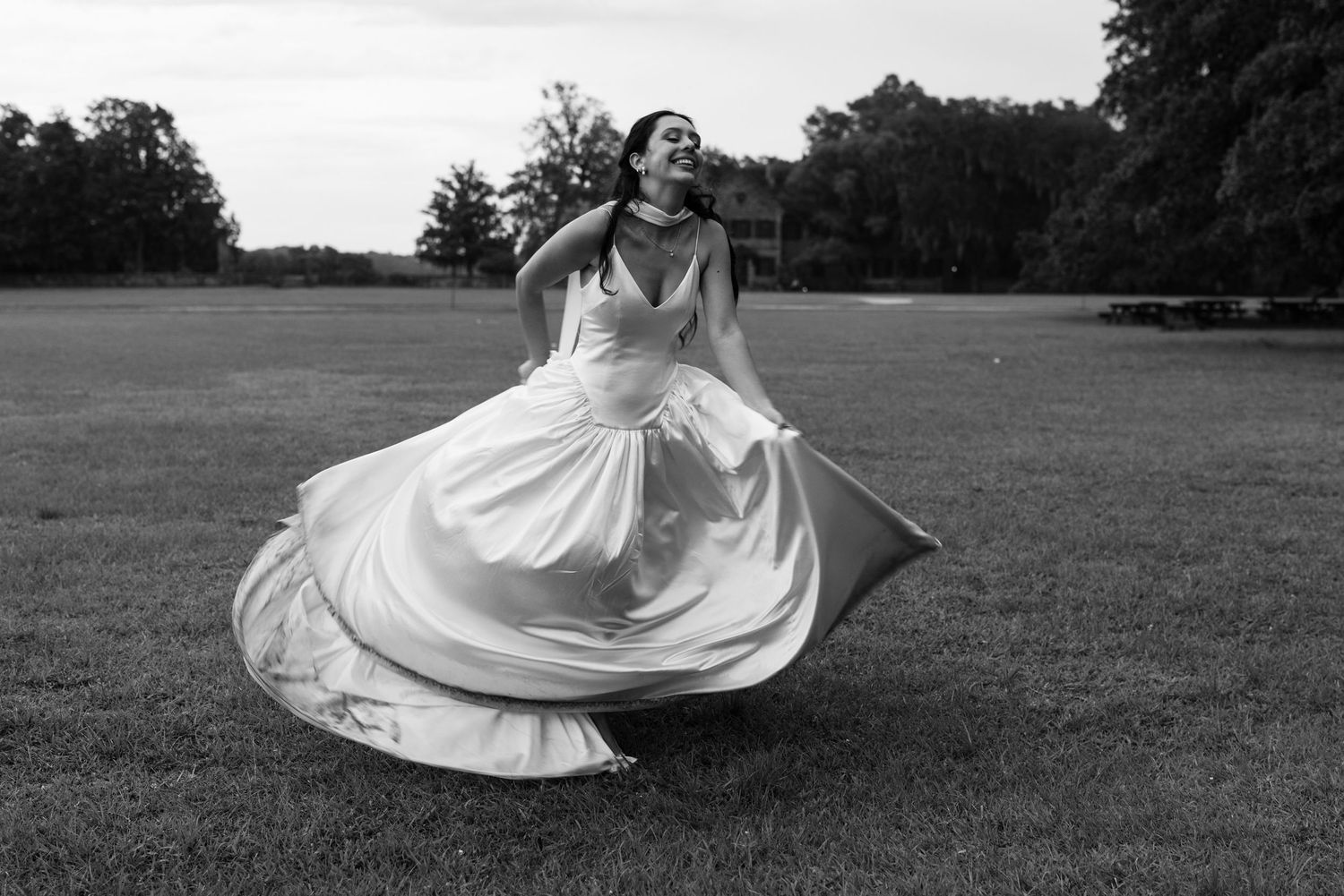 Black and white photo of joyful bride twirling in flowing satin wedding gown on open grass field.