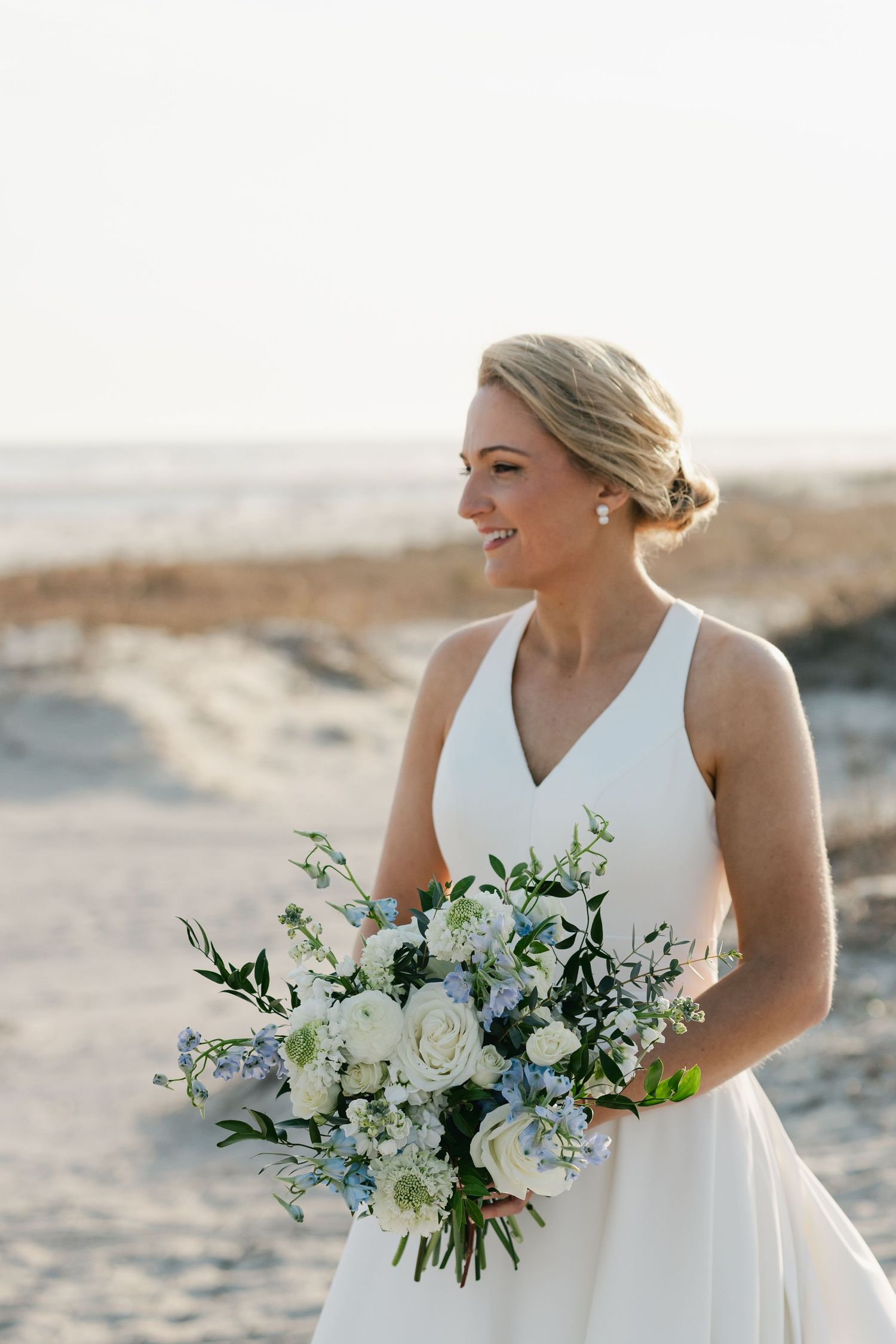 Smiling bride in white V-neck gown holds blue and white floral bouquet on sandy beach.