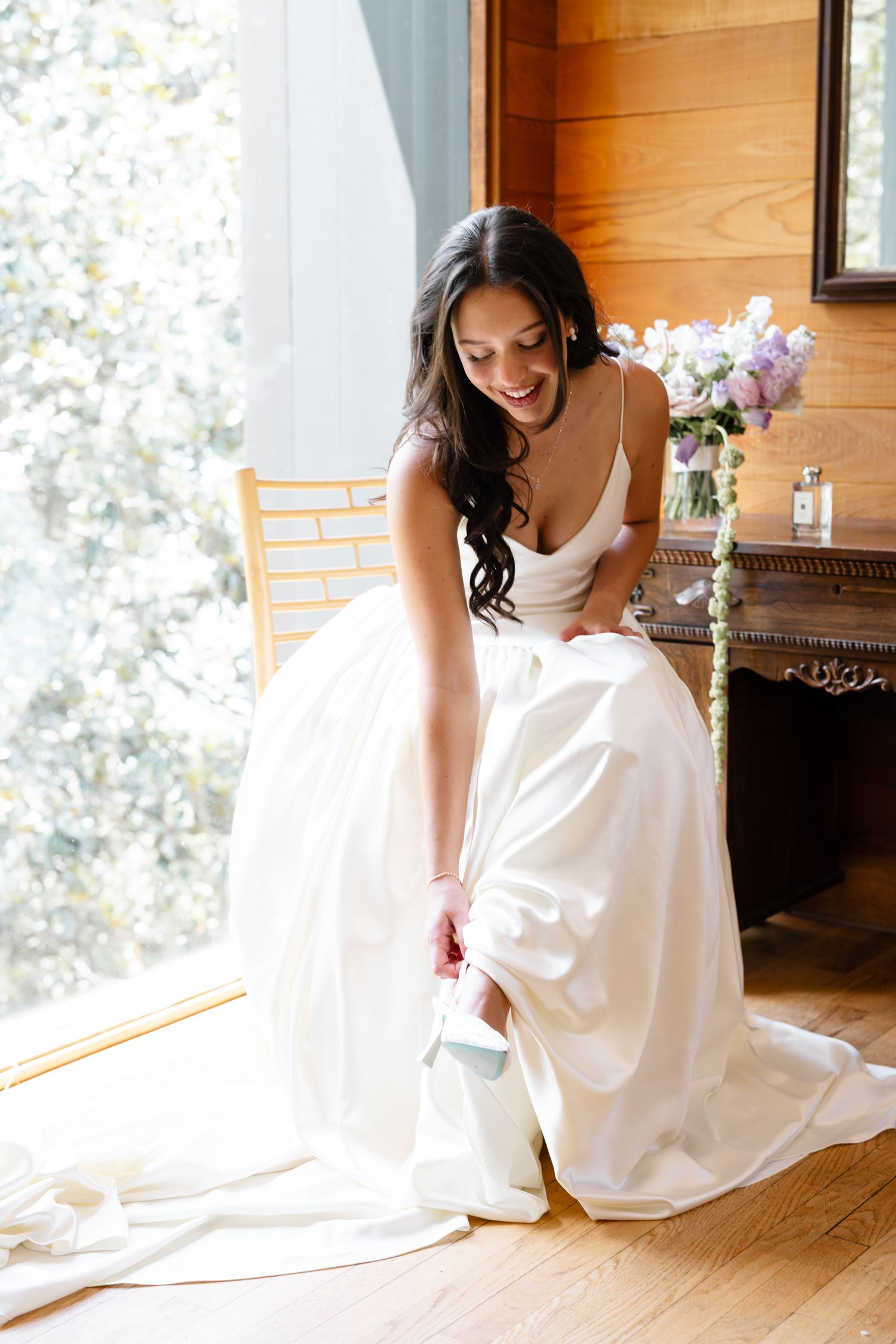 Smiling bride in white gown sitting on chair, adjusting her shoe, with floral arrangement in background.