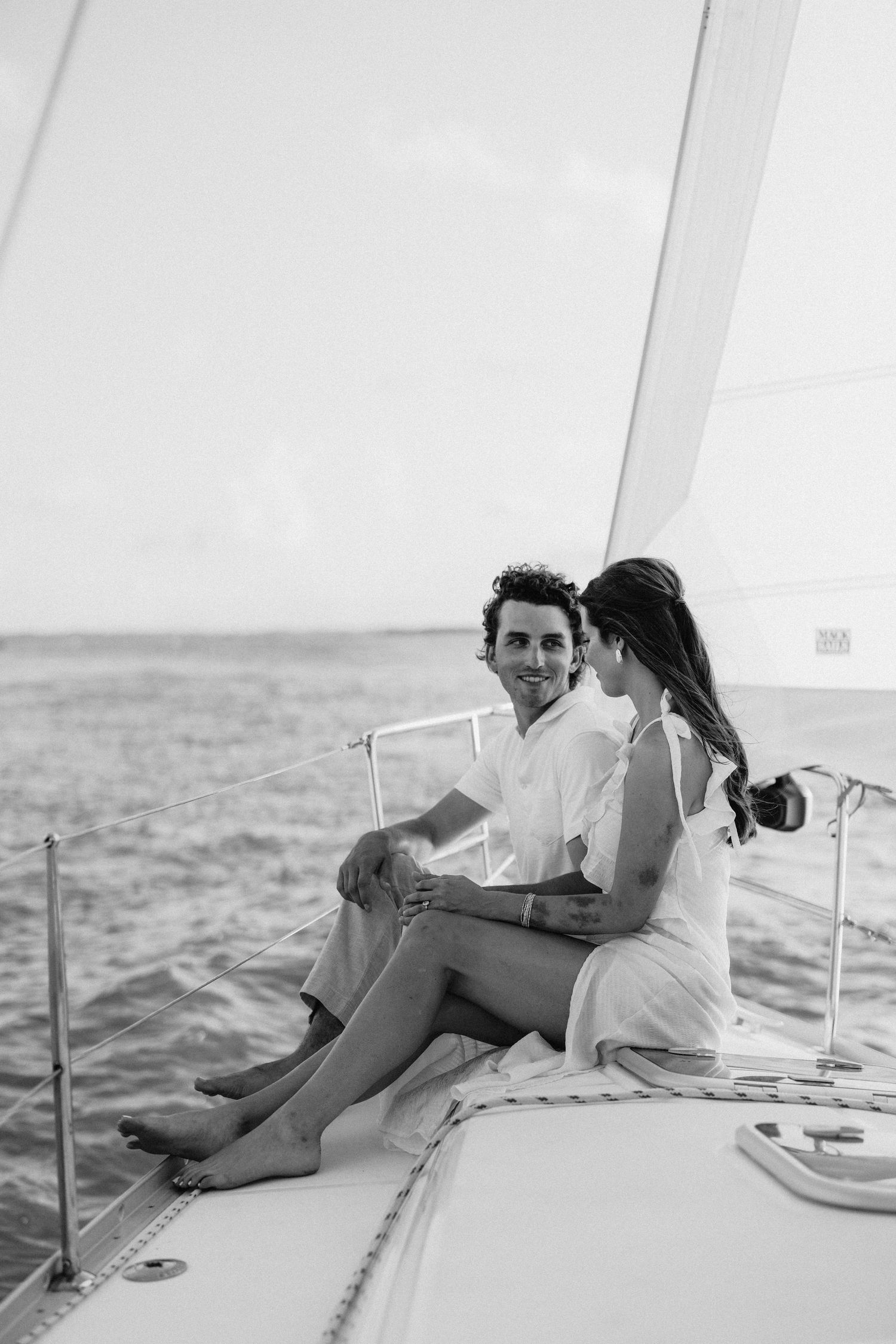 Black and white photo of a couple sitting together on a sailboat deck, smiling, with the ocean in the background.