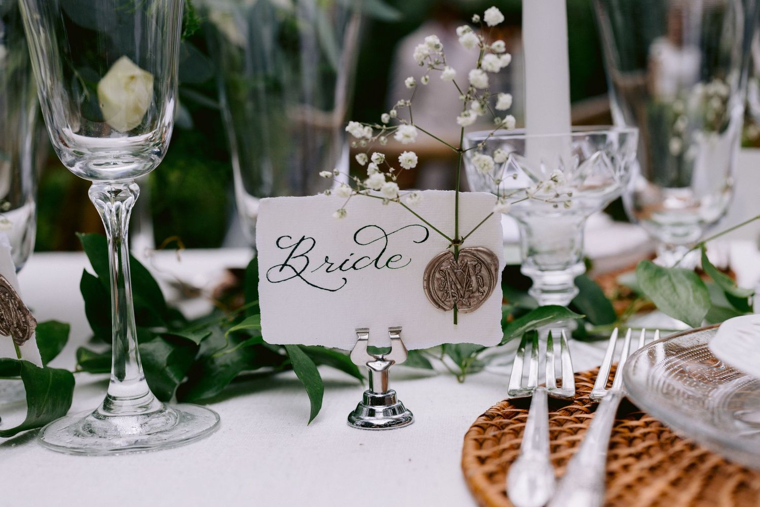 Elegant wedding reception table with calligraphy place card, crystal glasses, greenery, and candles.