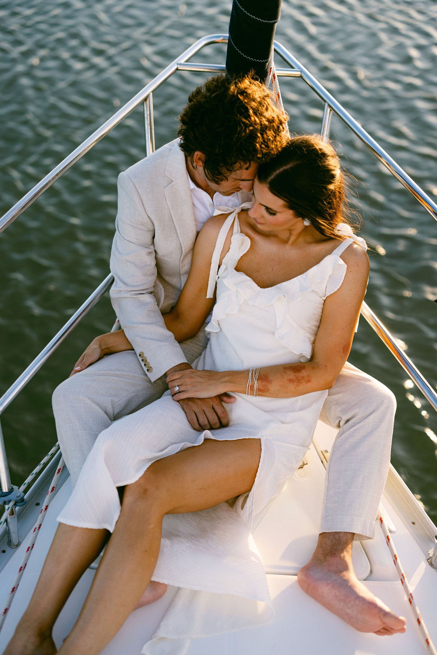 Couple sharing intimate moment on sailboat bow, dressed in white and beige, with sparkling water backdrop.
