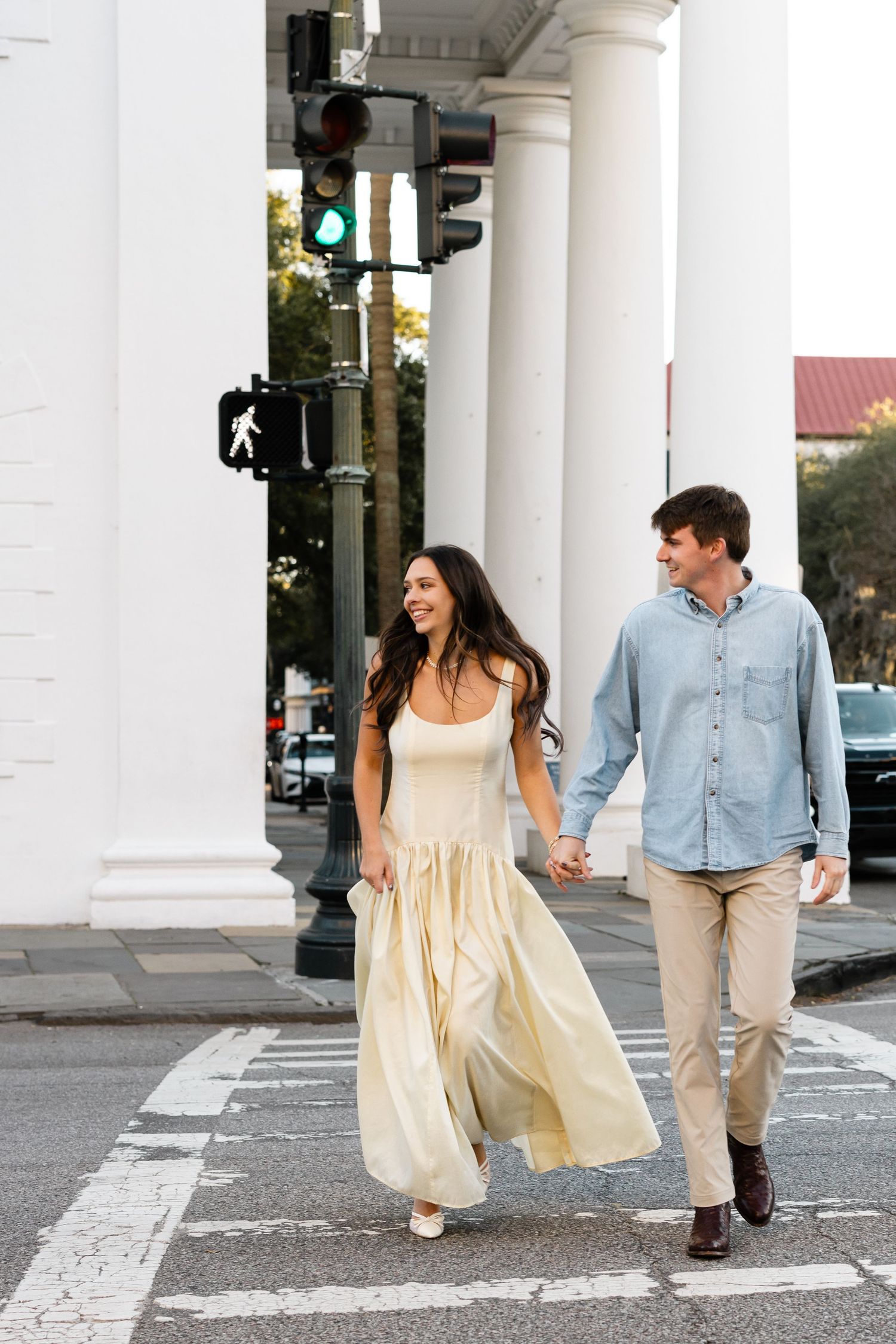 Couple holding hands crossing a city street, woman in yellow maxi dress, man in blue shirt and khaki pants.
