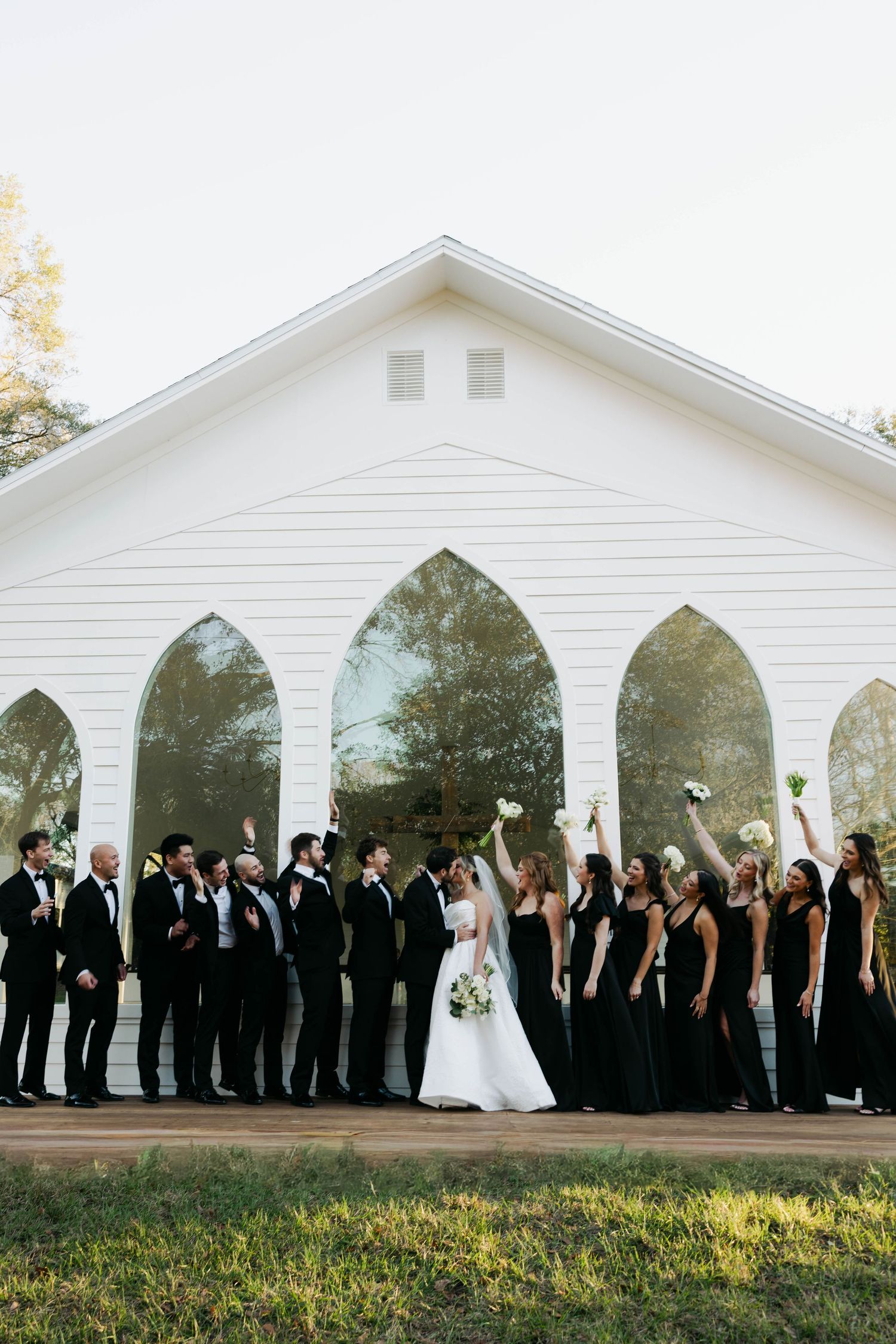 Wedding party in black attire cheering outside a white chapel church with gothic arch windows.