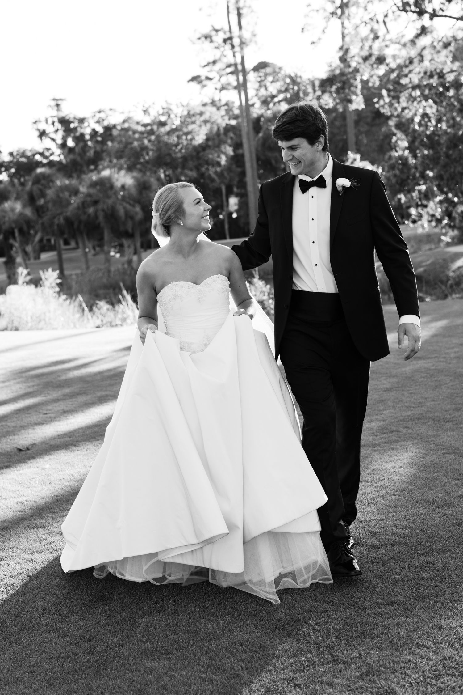 Black and white photo of a bride and groom walking together outdoors, smiling at each other on their wedding day.