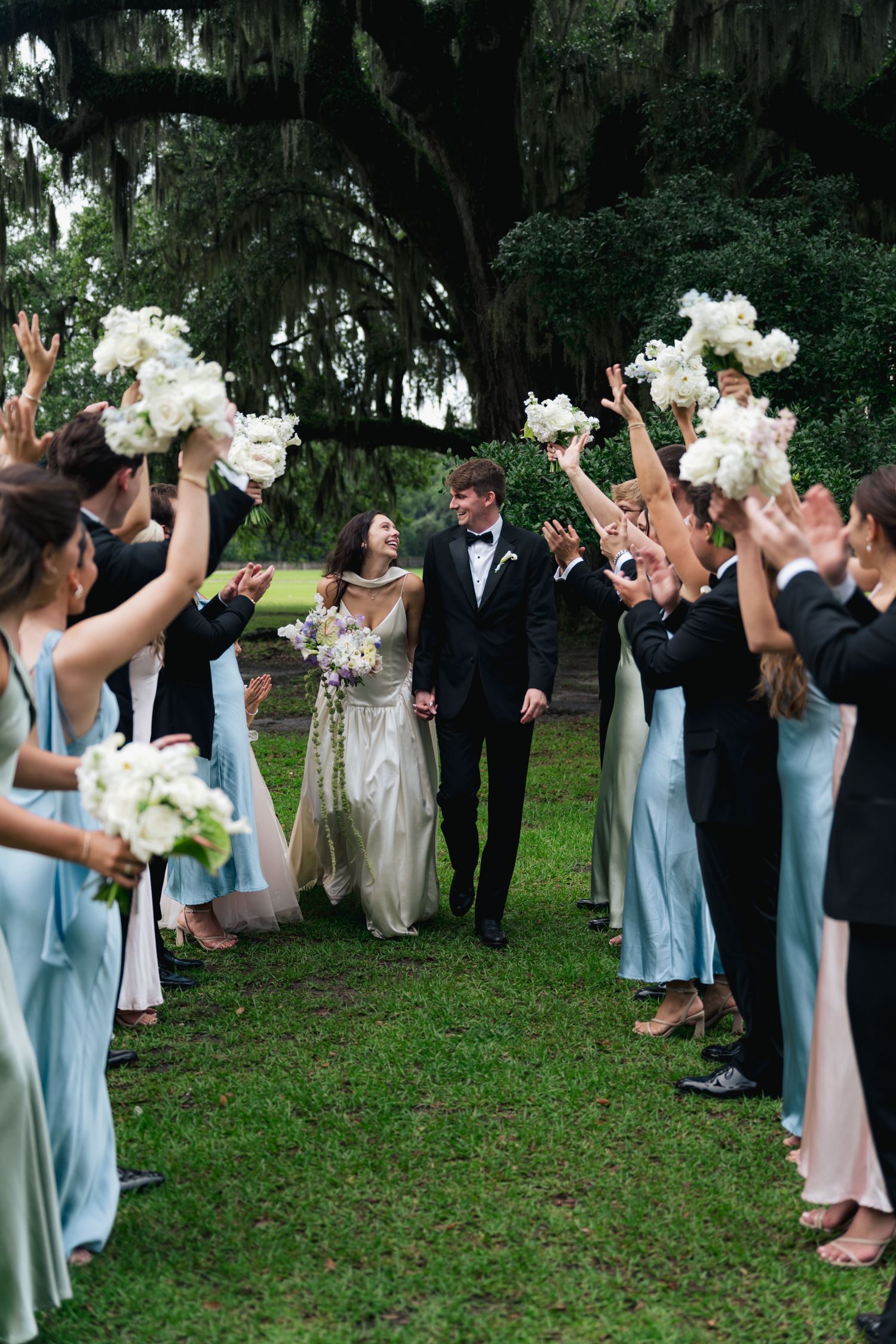 Bride and groom walk through cheering wedding party raising white bouquets under moss-draped oak trees.