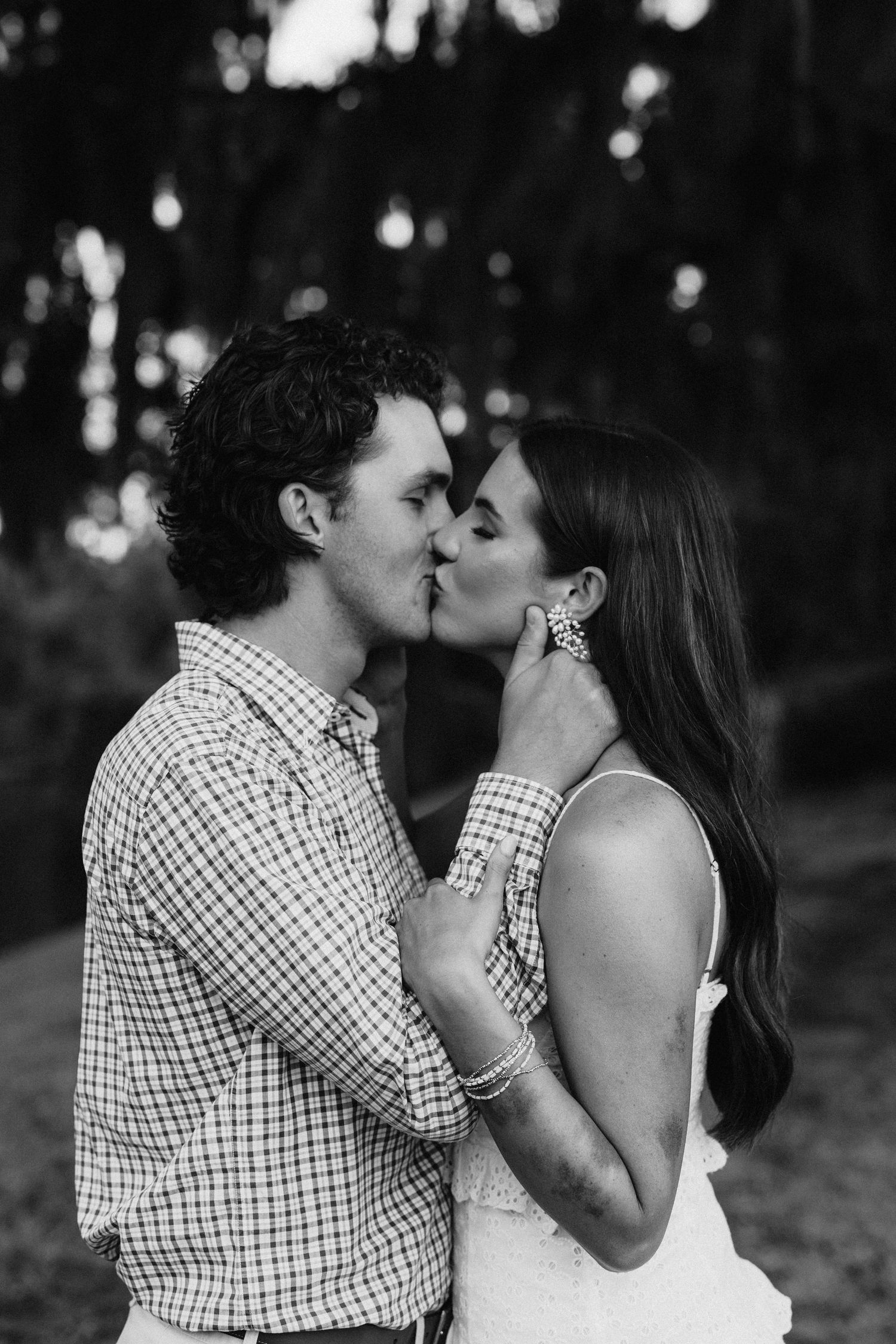 Black and white romantic photo of a couple sharing a tender kiss outdoors in a wooded setting.