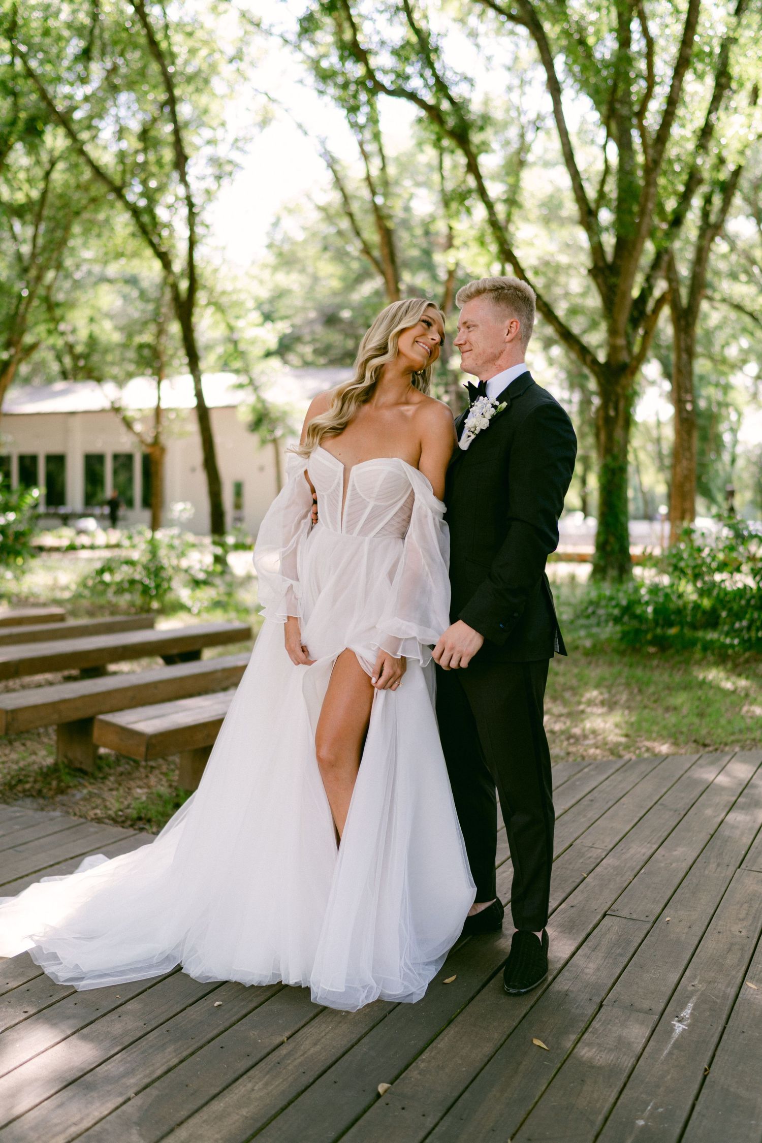 Smiling bride in off-shoulder white gown with leg slit and groom in black tux share tender outdoor moment.