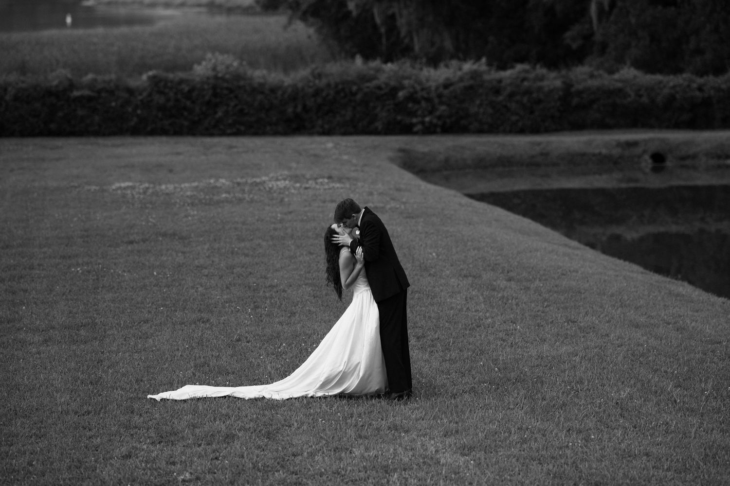 Black and white romantic photo of bride and groom kissing on vast green lawn with long flowing train.