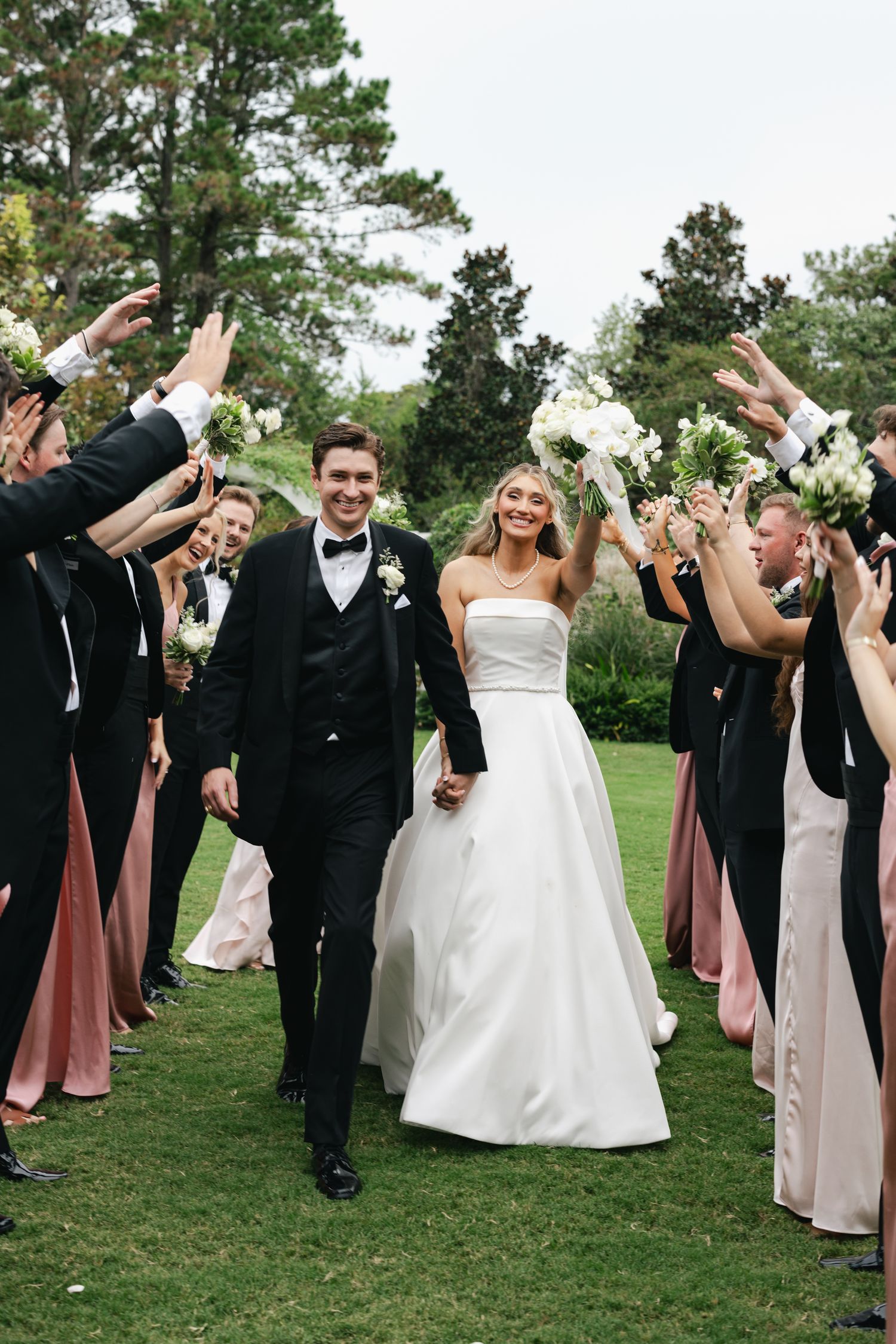 Bride and groom walk hand-in-hand through a wedding party tunnel on a green lawn, surrounded by cheering guests.