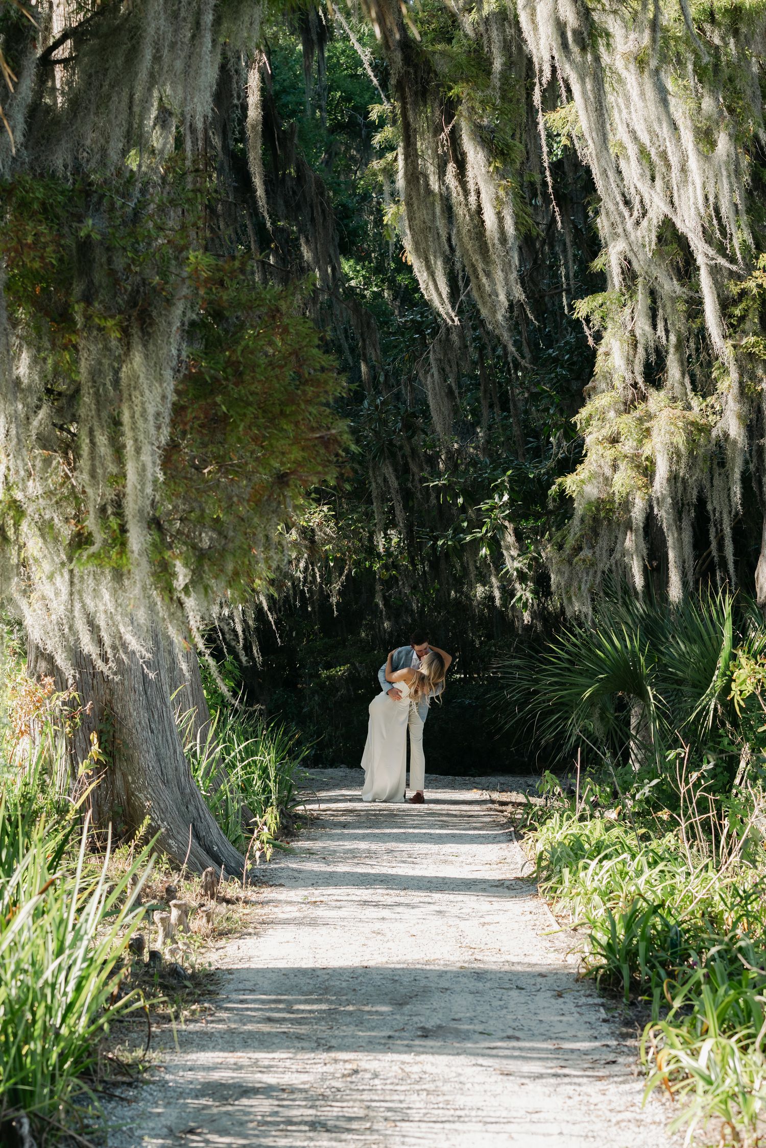 Bride and groom embrace on a tree-lined path draped with Spanish moss in a lush garden setting.