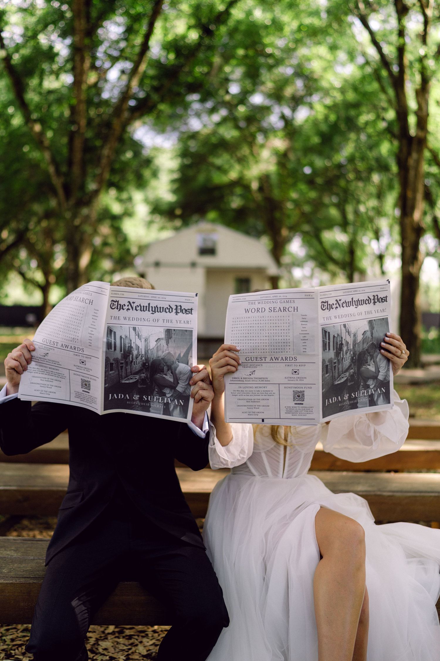 Bride and groom sitting on a bench hiding their faces behind custom wedding newspapers outdoors.