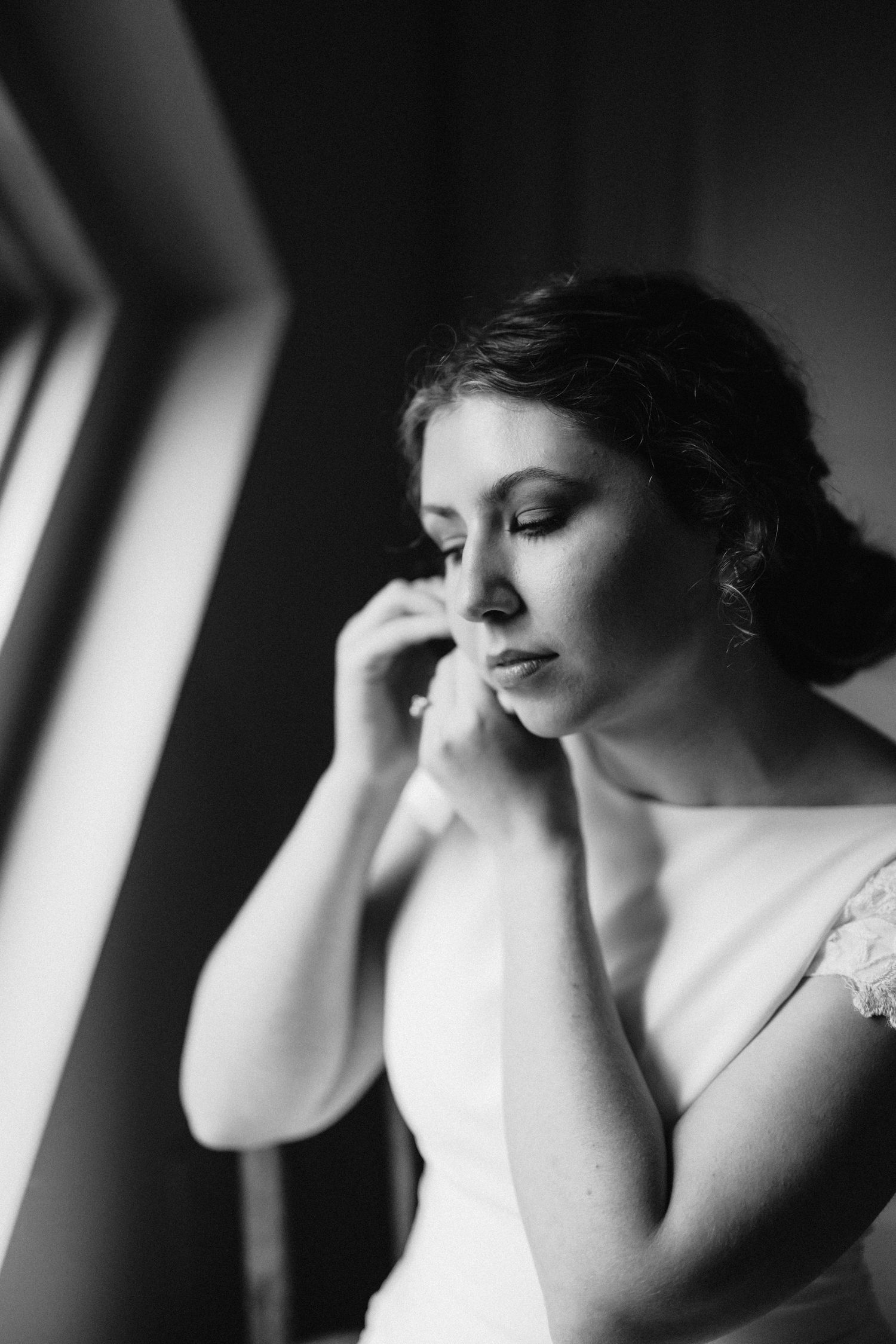 Black and white portrait of bride in elegant gown adjusting earring beside soft window light.