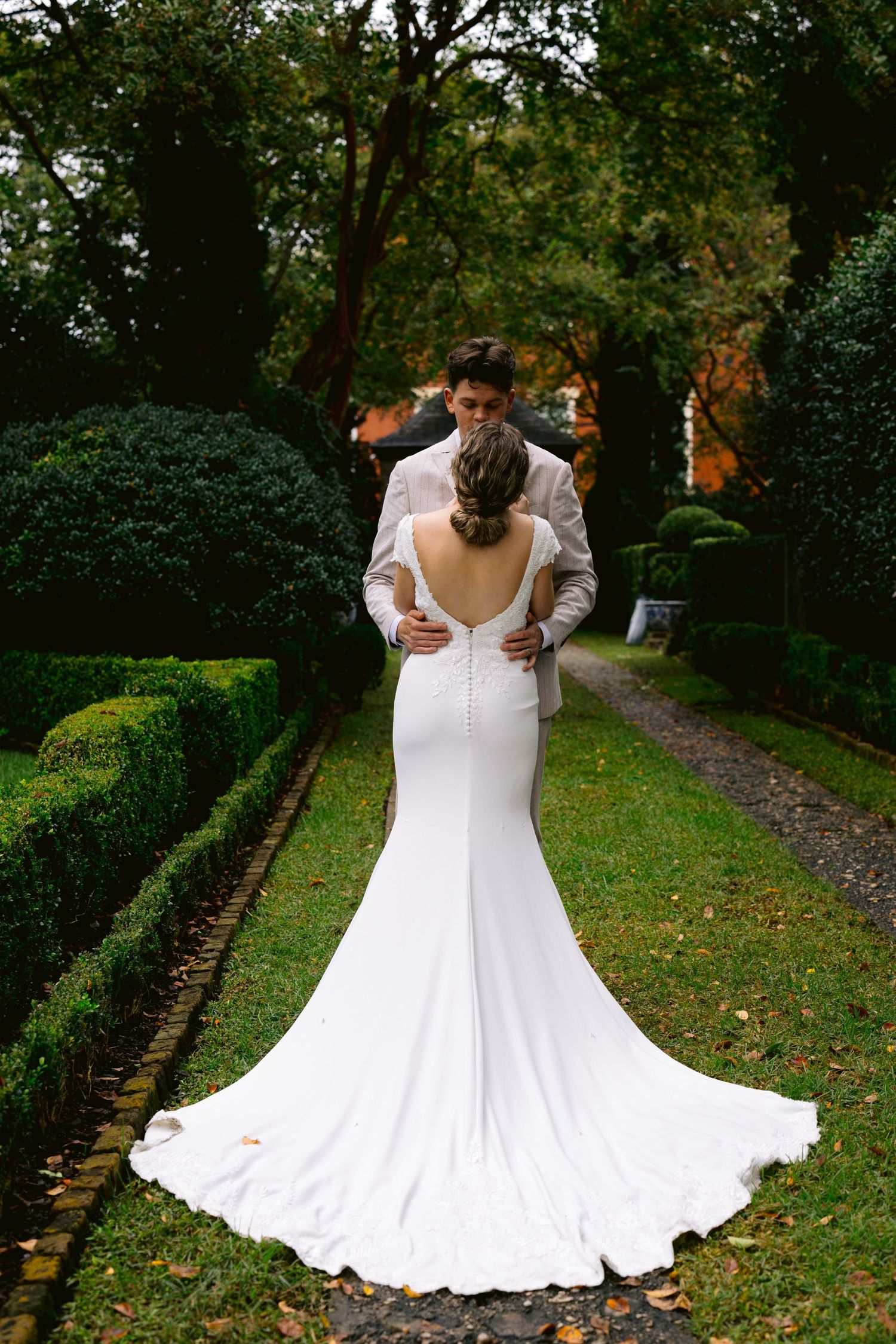Rear view of bride in fitted white gown with button back and long train in formal garden path.