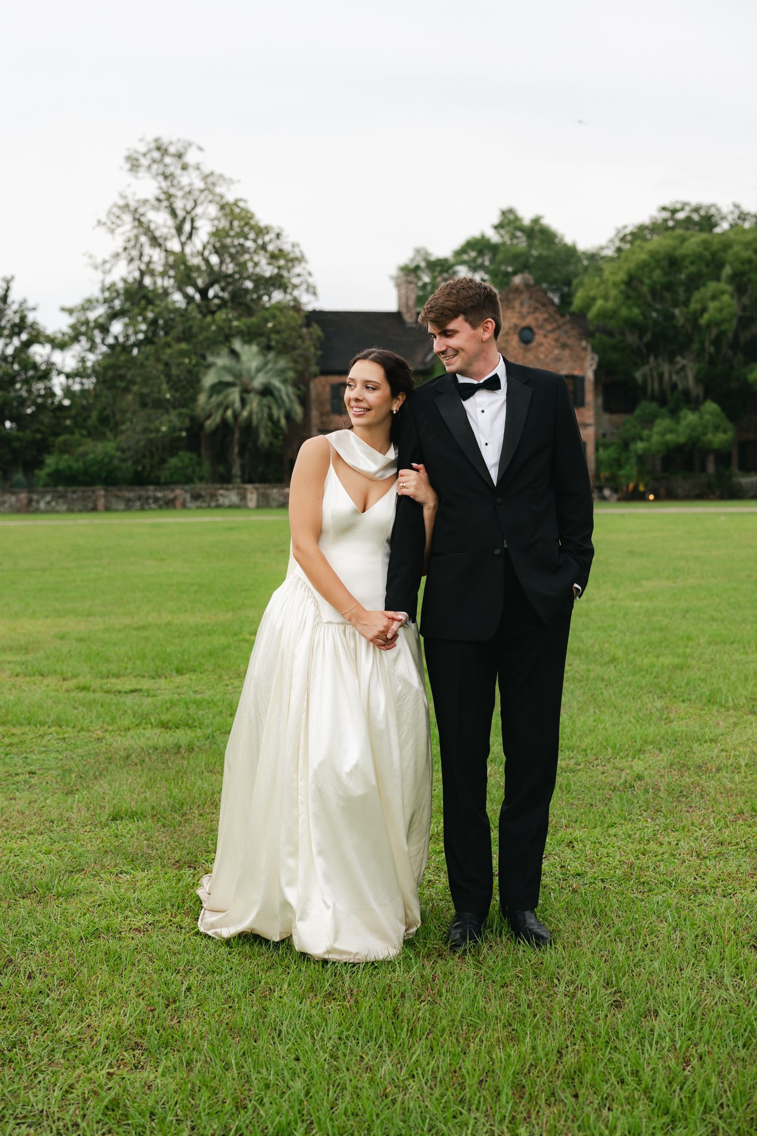 Bride in white gown and groom in black tuxedo stand together on green lawn with historic brick building backdrop.