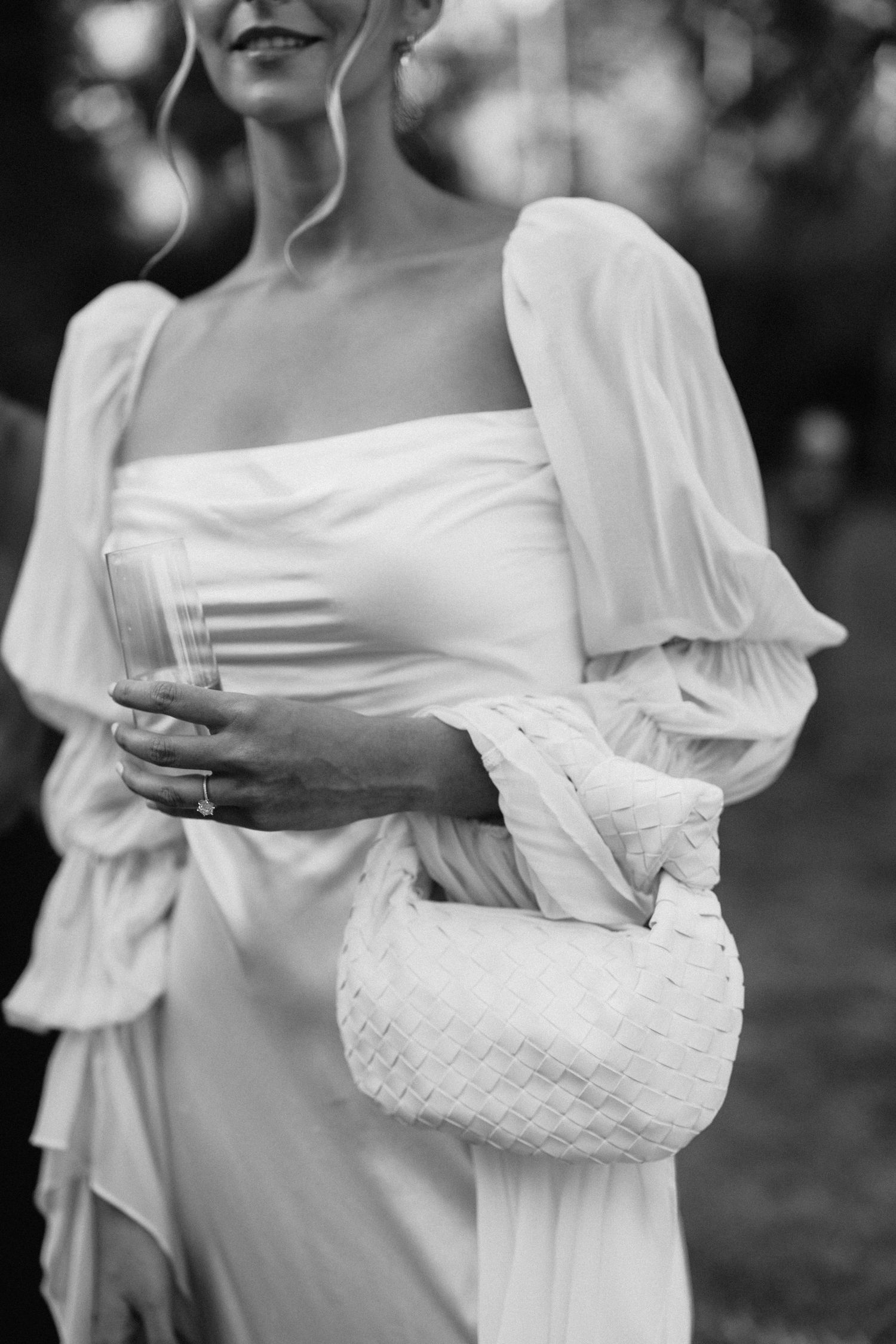 Bride in white off-shoulder gown with billowy sleeves holding a woven white clutch bag, black and white photo.